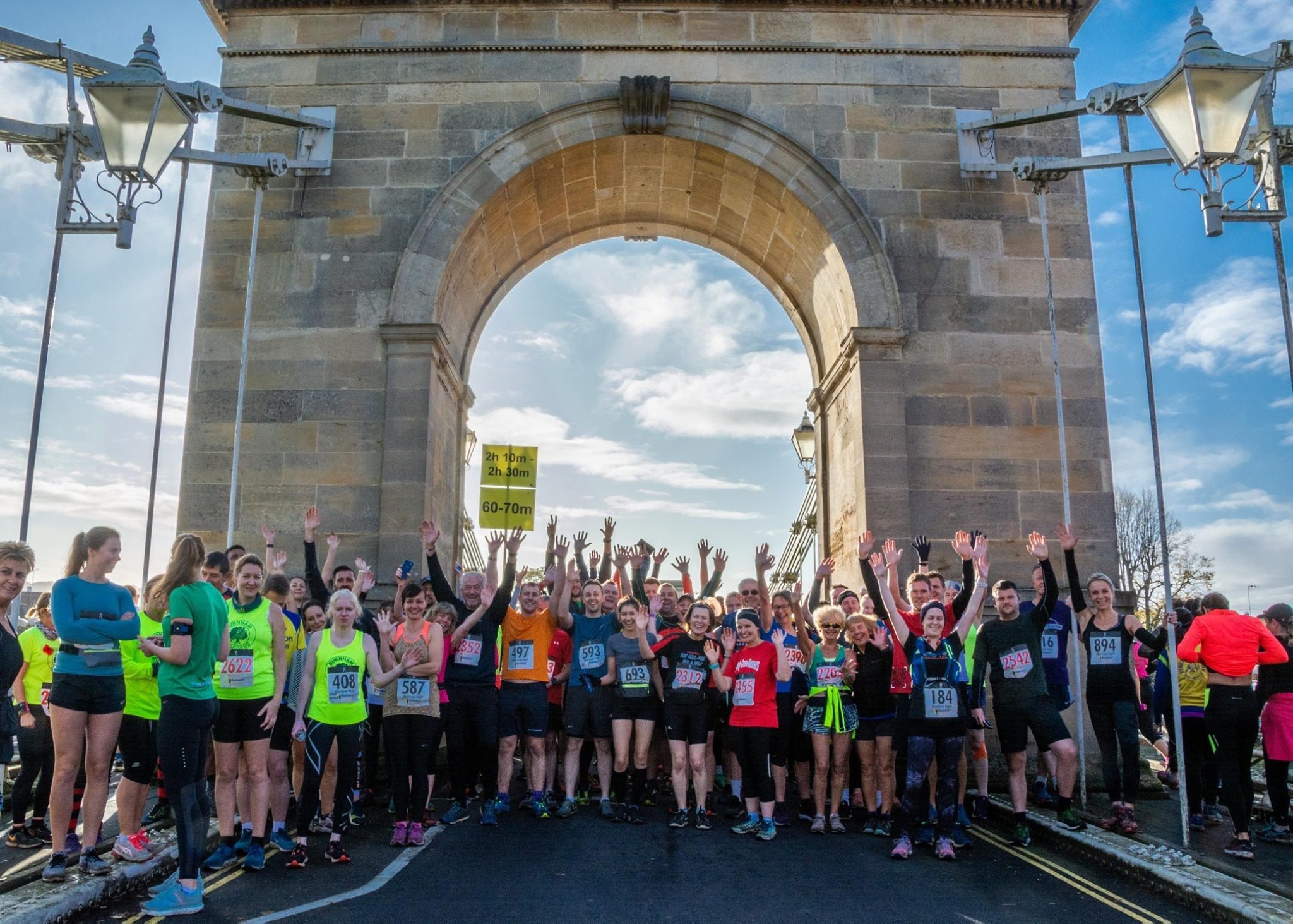 A large group of runners stand together with their arms raised at the starting line of a race, under a stone archway. They are dressed in athletic gear and numbered bibs, and the sky above is partly cloudy with visible patches of blue.