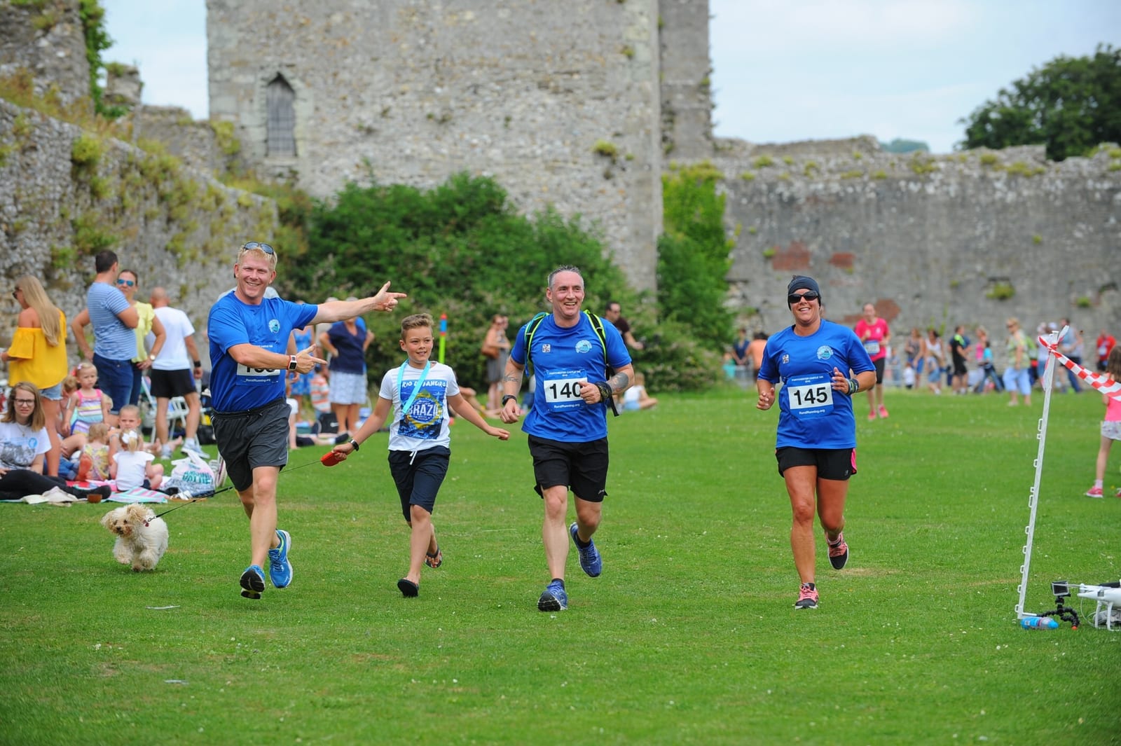Four runners in blue shirts, three men and one woman, approaching the finish line in a grassy area with stone ruins in the background. Onlookers sit on the grass, and a small dog is seen in the foreground. The runners are smiling and appear to be joyful.