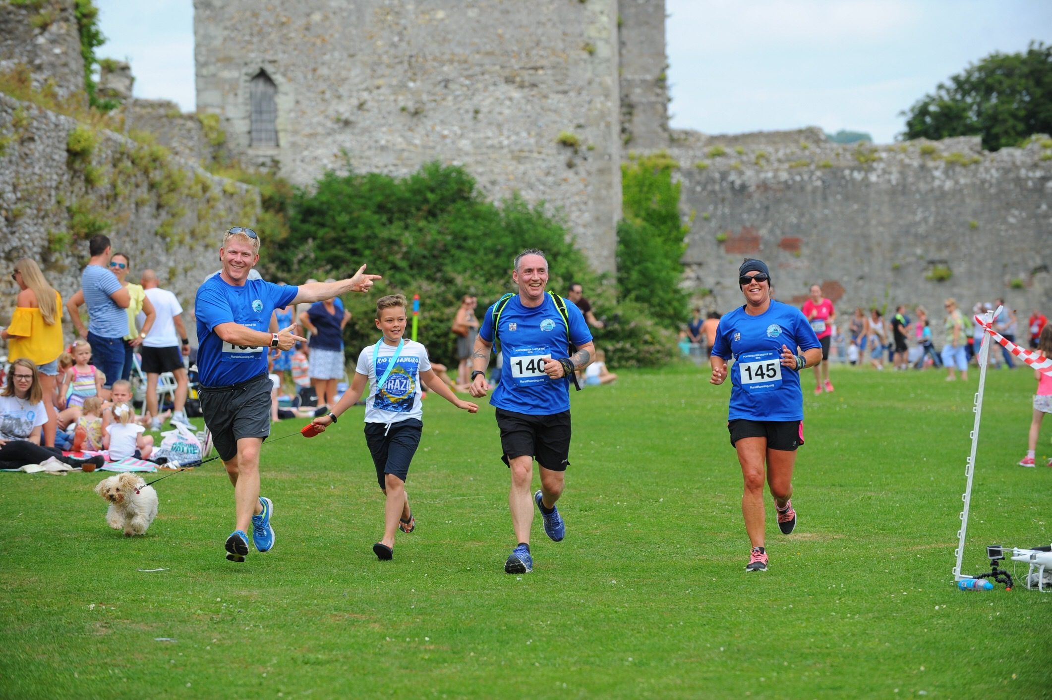Four runners in blue shirts, three men and one woman, approaching the finish line in a grassy area with stone ruins in the background. Onlookers sit on the grass, and a small dog is seen in the foreground. The runners are smiling and appear to be joyful.