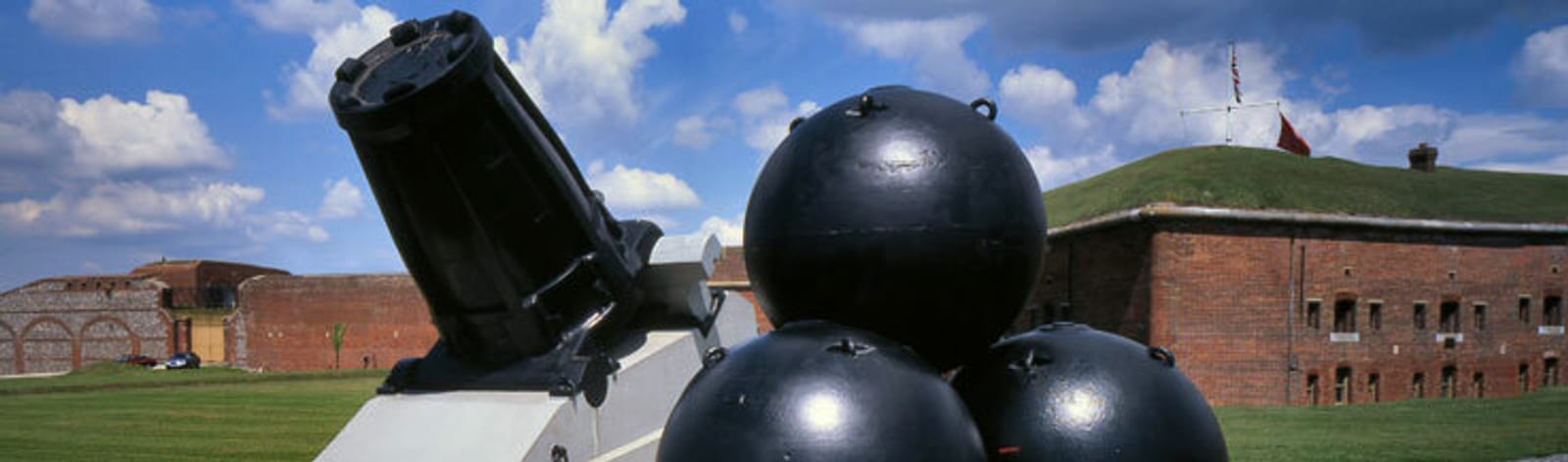 A historical fort with red brick walls is visible under a partly cloudy sky. In the foreground, there are several large black cannonballs and part of a cannon. Green grass surrounds the area, and a flag is seen atop the fort in the background.