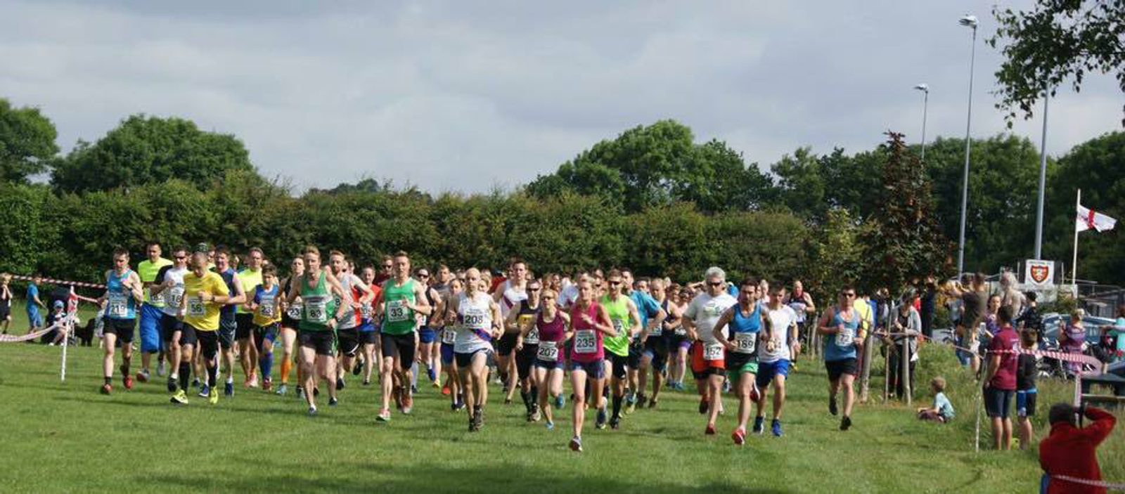 A large group of runners at the start of a race on a grassy field, each wearing a numbered bib. The runners are dressed in a variety of athletic clothing. Spectators are seen in the background, along with trees and a cloudy sky.