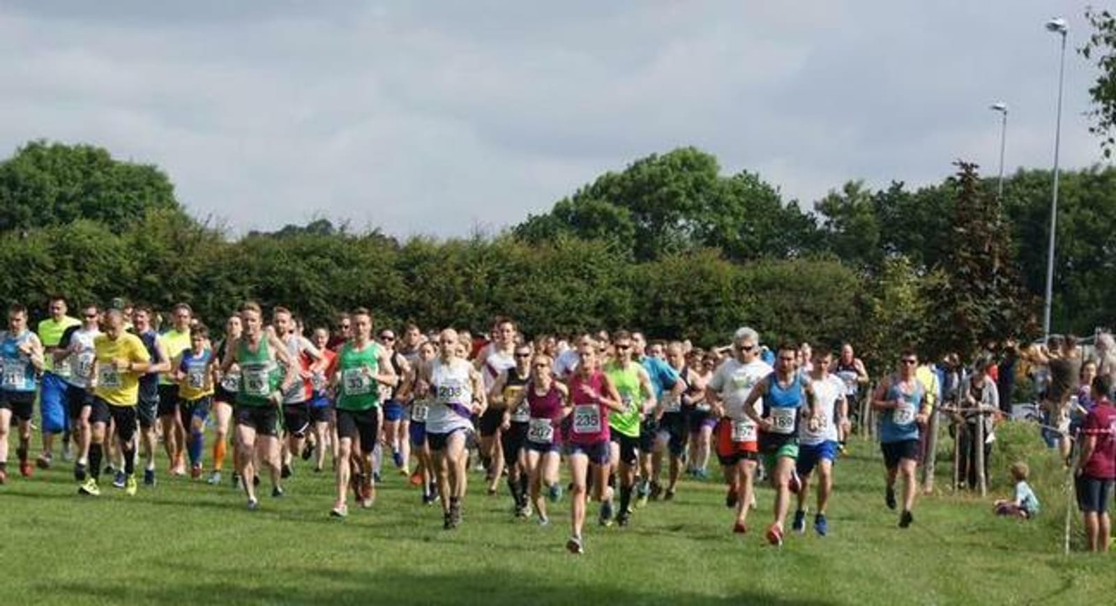 A large group of runners wearing colorful athletic gear are participating in an outdoor race on a grassy field. They are running toward the camera, with spectators in the background. The sky is cloudy, and there are trees and bushes lining the field.