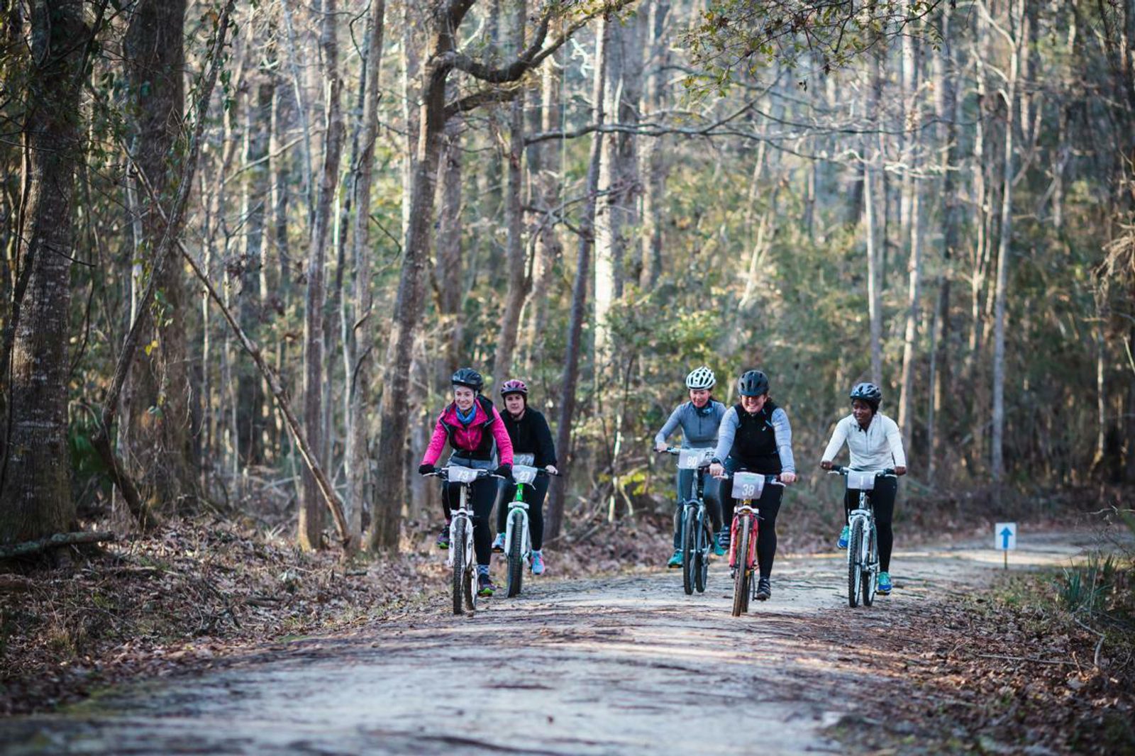 A group of five cyclists rides along a forest trail surrounded by tall, leafless trees. The cyclists, dressed in various colorful jackets and helmets, appear to be enjoying a sunny day outdoors. The ground is covered with dry leaves, and the scene is serene and natural.