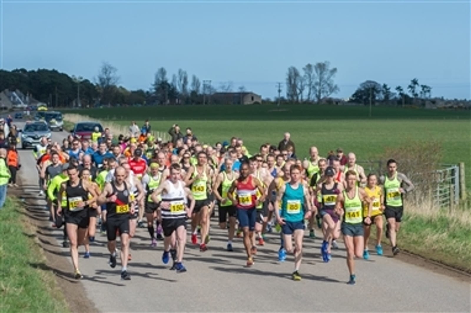 A large group of runners, wearing colorful athletic attire and race numbers, participate in an outdoor road race on a sunny day. The race takes place on a rural road with farmland and trees visible in the background.