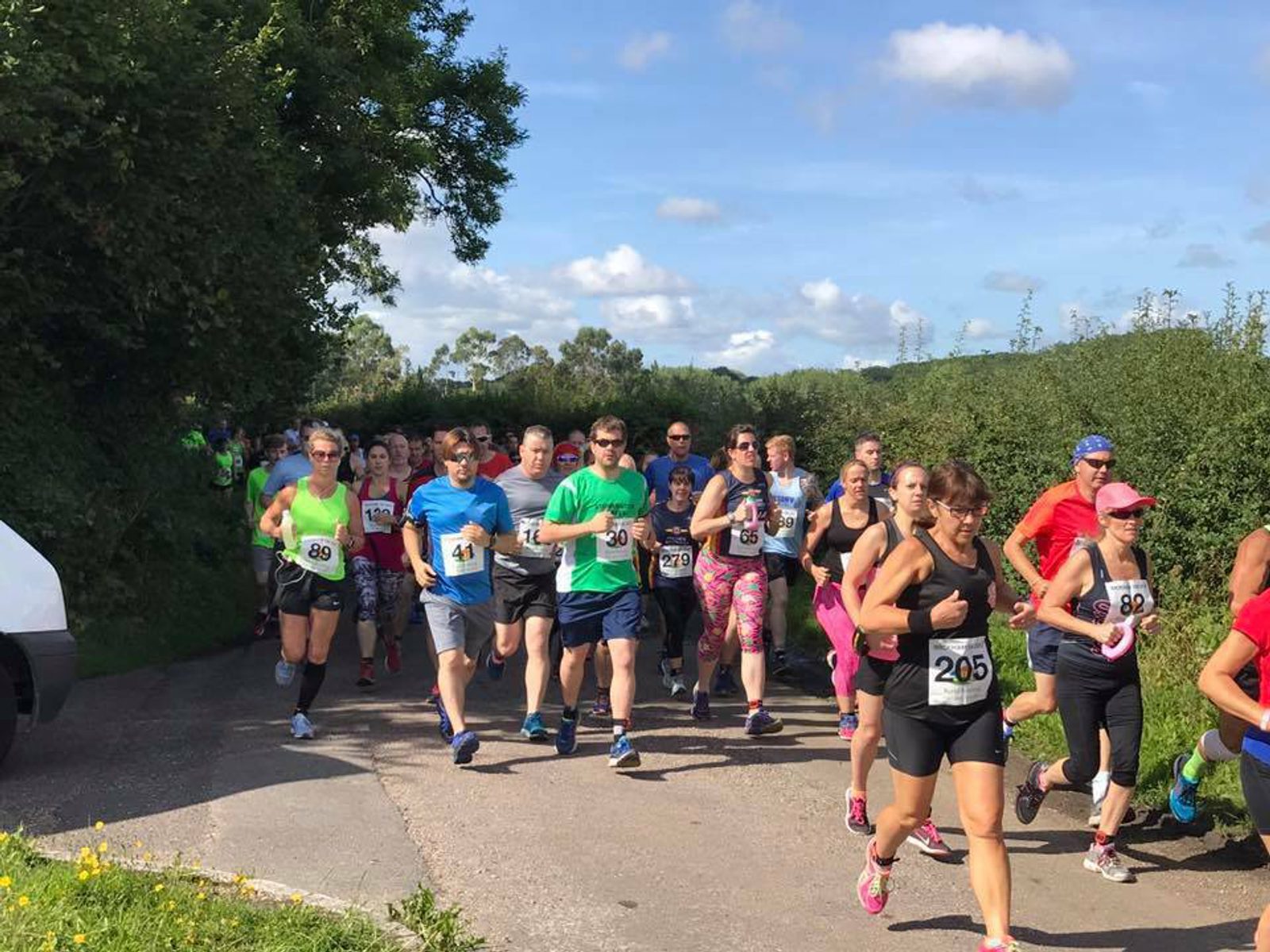 A group of runners, wearing various colorful outfits and displaying race numbers, participates in a race on a sunny day. They are running on a country road surrounded by greenery, with a few spectators and a parked white van visible in the background.