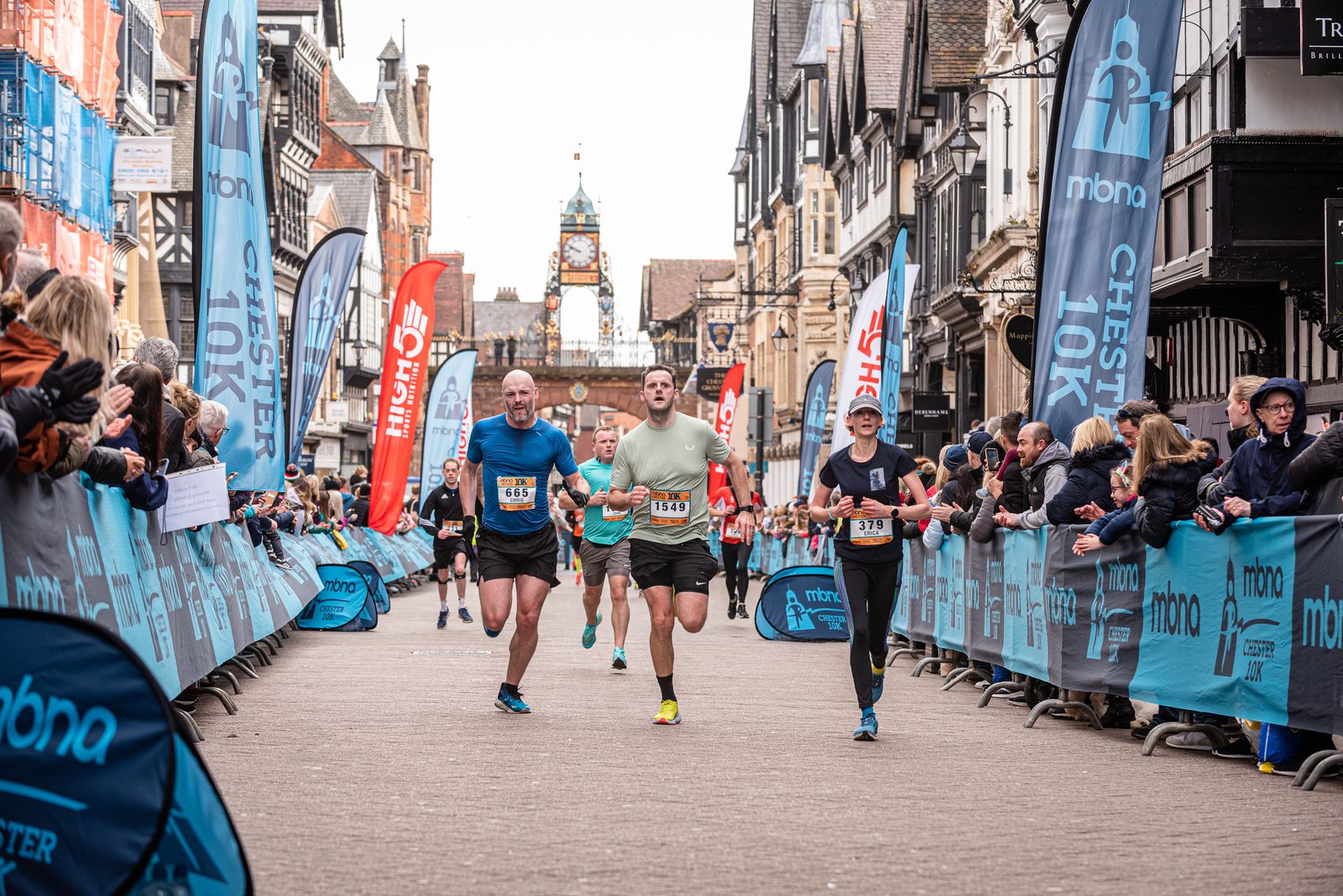 Runners approach the finish line in a crowded street race with cheering spectators on both sides. Flags and banners display the event's logo, and historic buildings line the street. The atmosphere is lively and celebratory.