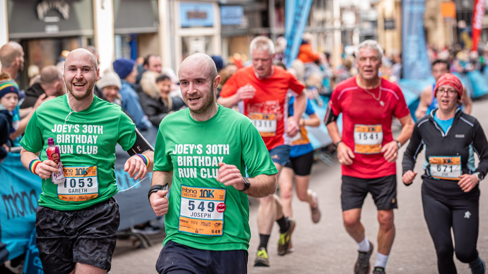A group of runners in a road race, including two men in green "Joey's 30th Birthday Run Club" shirts. The leading man wears bib 549, smiling. The participants behind them wear various colorful sportswear. Spectators line the street, cheering the runners on.