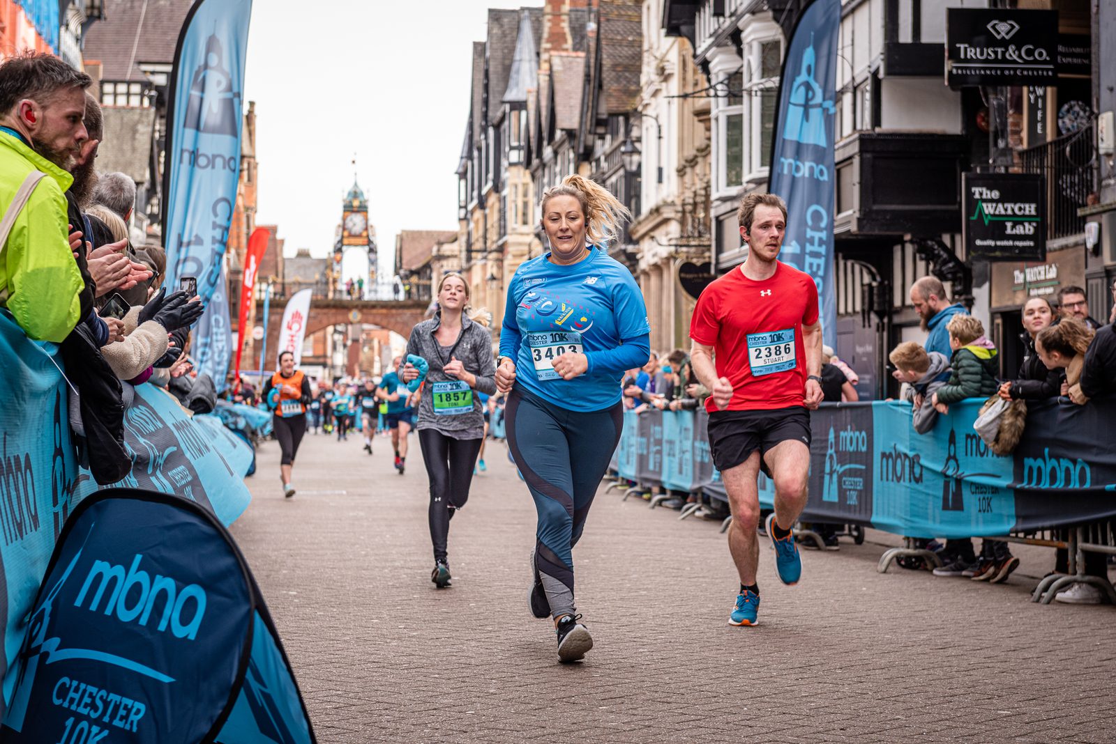 Runners participating in a race on a city street lined with spectators. Banners and barriers are seen along the path. A woman in a blue top runs in the center, a man in a red top and a woman in a green top run nearby. Buildings and a clock tower are visible in the background.