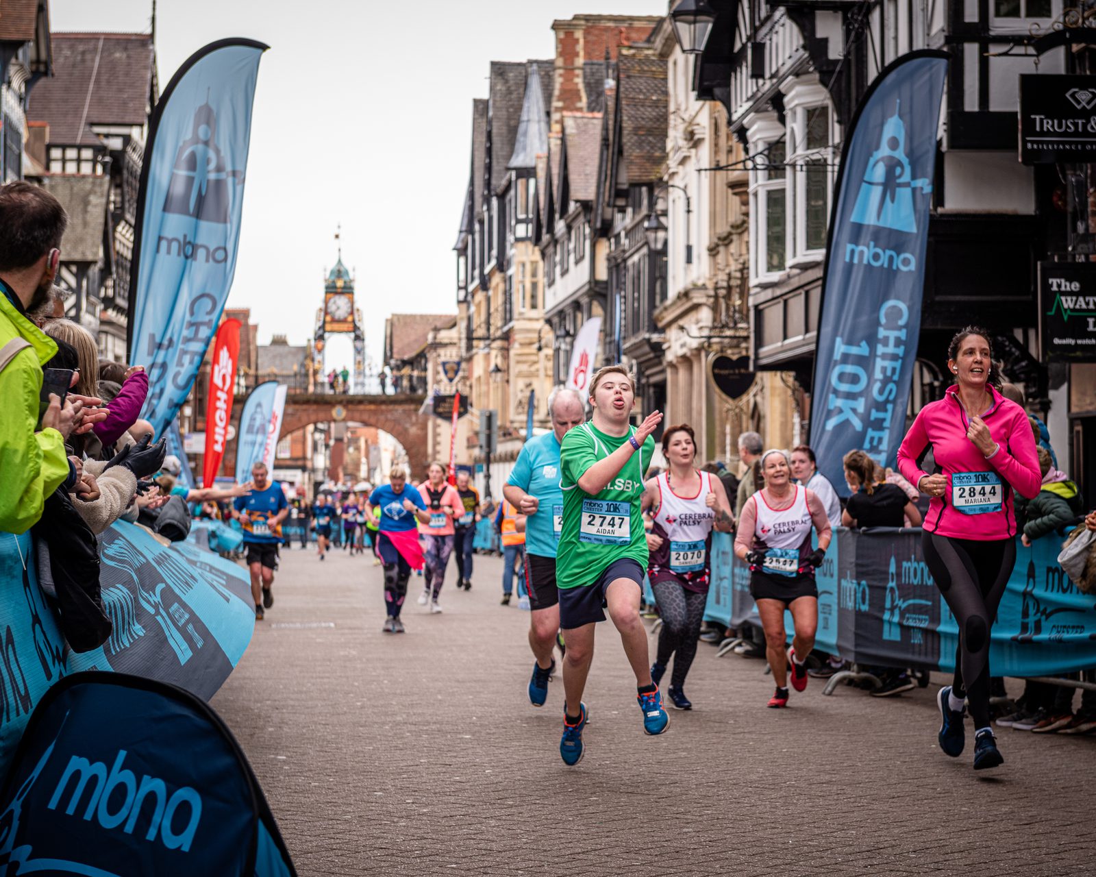 Runners participating in a 10K race fill a street lined with cheering spectators. The backdrop features historic buildings and a clock tower. Participants wear numbered bibs, with some looking determined and others smiling. Event banners and flags are visible along the route.