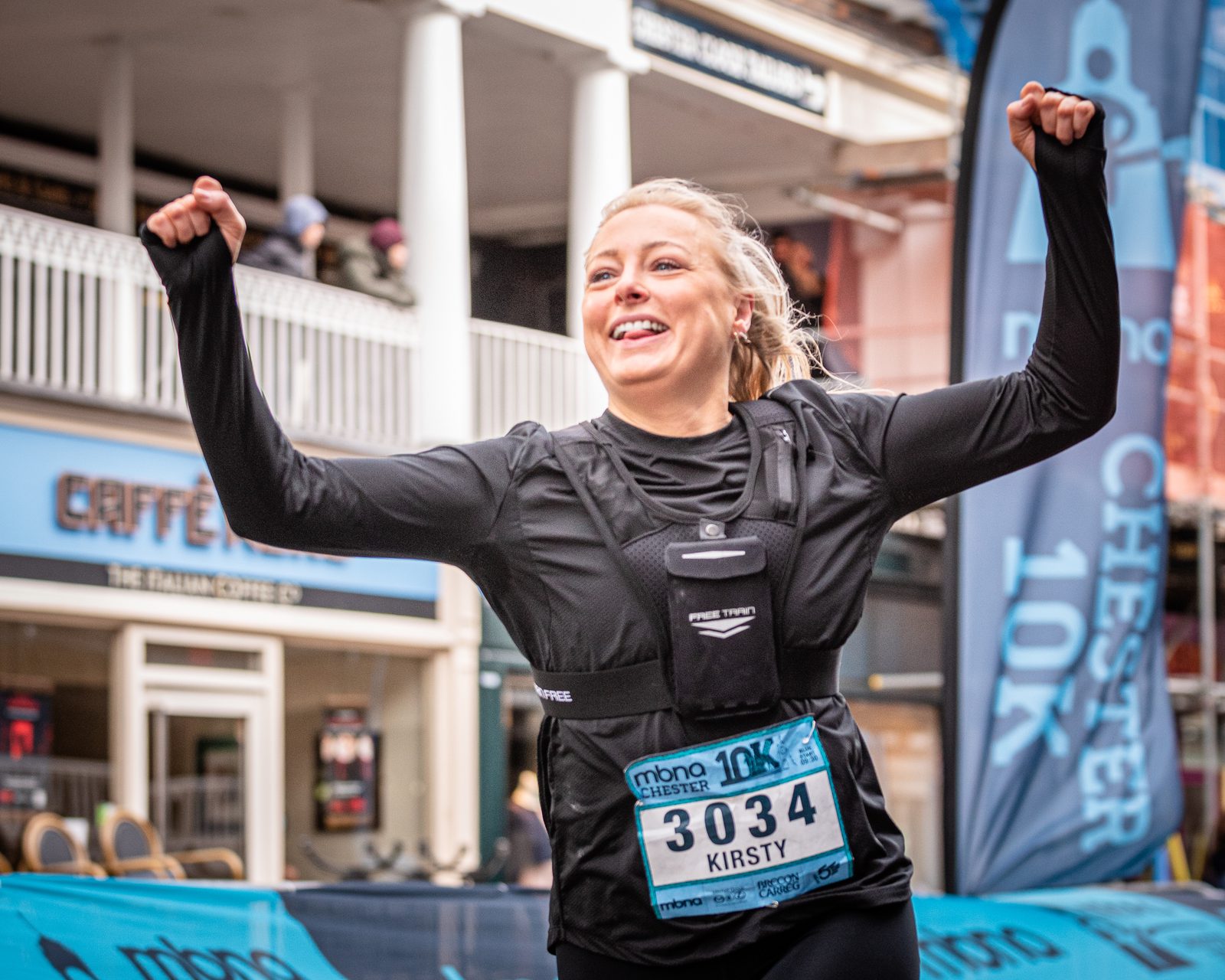 A female runner, wearing a race bib numbered 3034 and named Kirsty, celebrates by raising her arms in victory. She is smiling widely as she crosses the finish line of the Chester 10K race, evidenced by the "Chester 10K" event branding visible in the background.