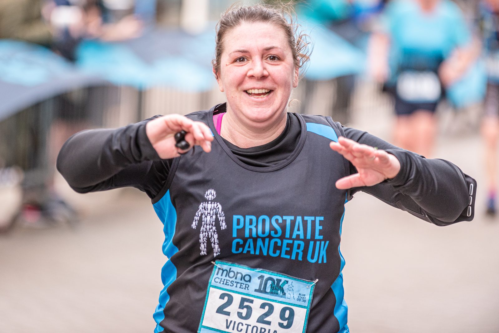 A runner wearing a "Prostate Cancer UK" shirt, with a race bib numbered 2529 and named Victoria, smiles as she nears the finish line of a 10K race. The background shows blurry spectators and other participants.