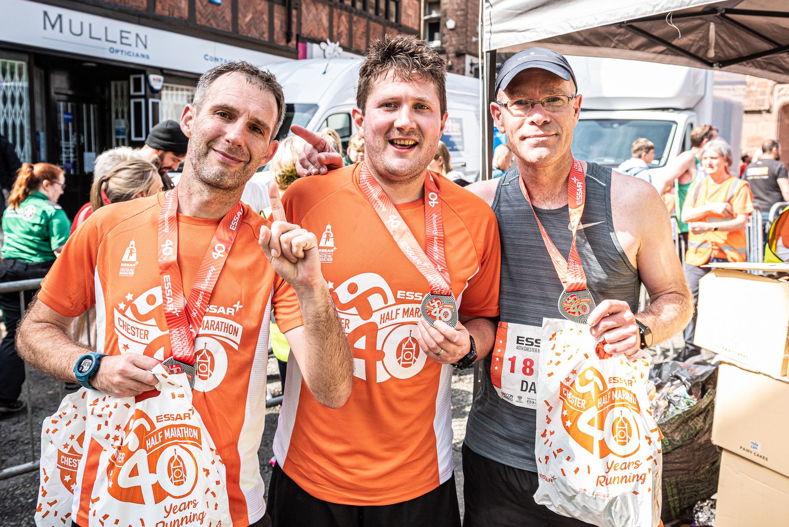 Three men proudly display their finisher medals and event bags after completing a half marathon. They are wearing orange race shirts and appear sweaty and tired but happy. Behind them, other participants and event staff can be seen, along with a white tent and a storefront.