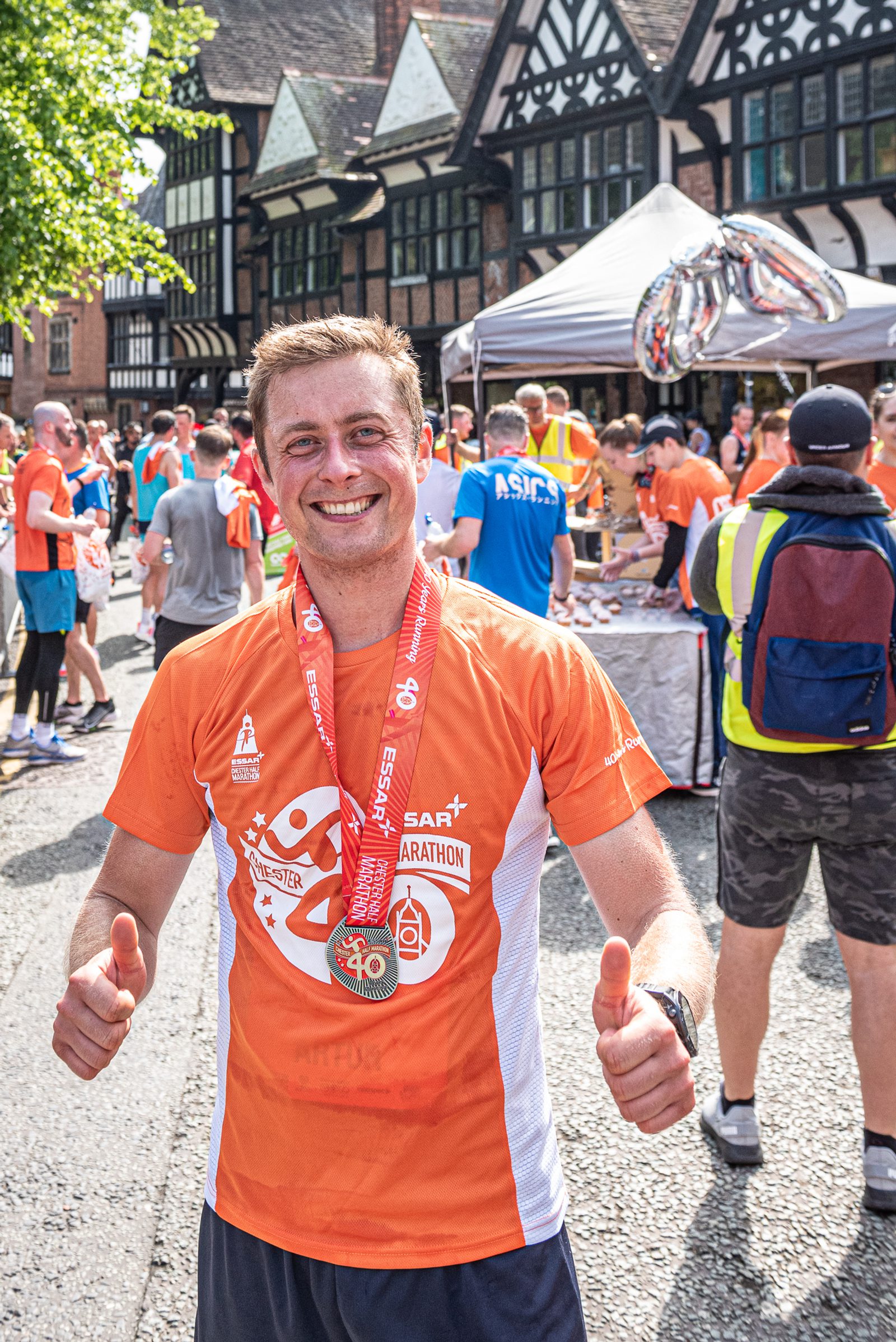 A man wearing an orange shirt and a marathon medal stands smiling with both thumbs up. He is among a group of people outdoors, some wearing similar shirts and a volunteer in a neon vest. Tudor-style buildings and a canopy are visible in the background.