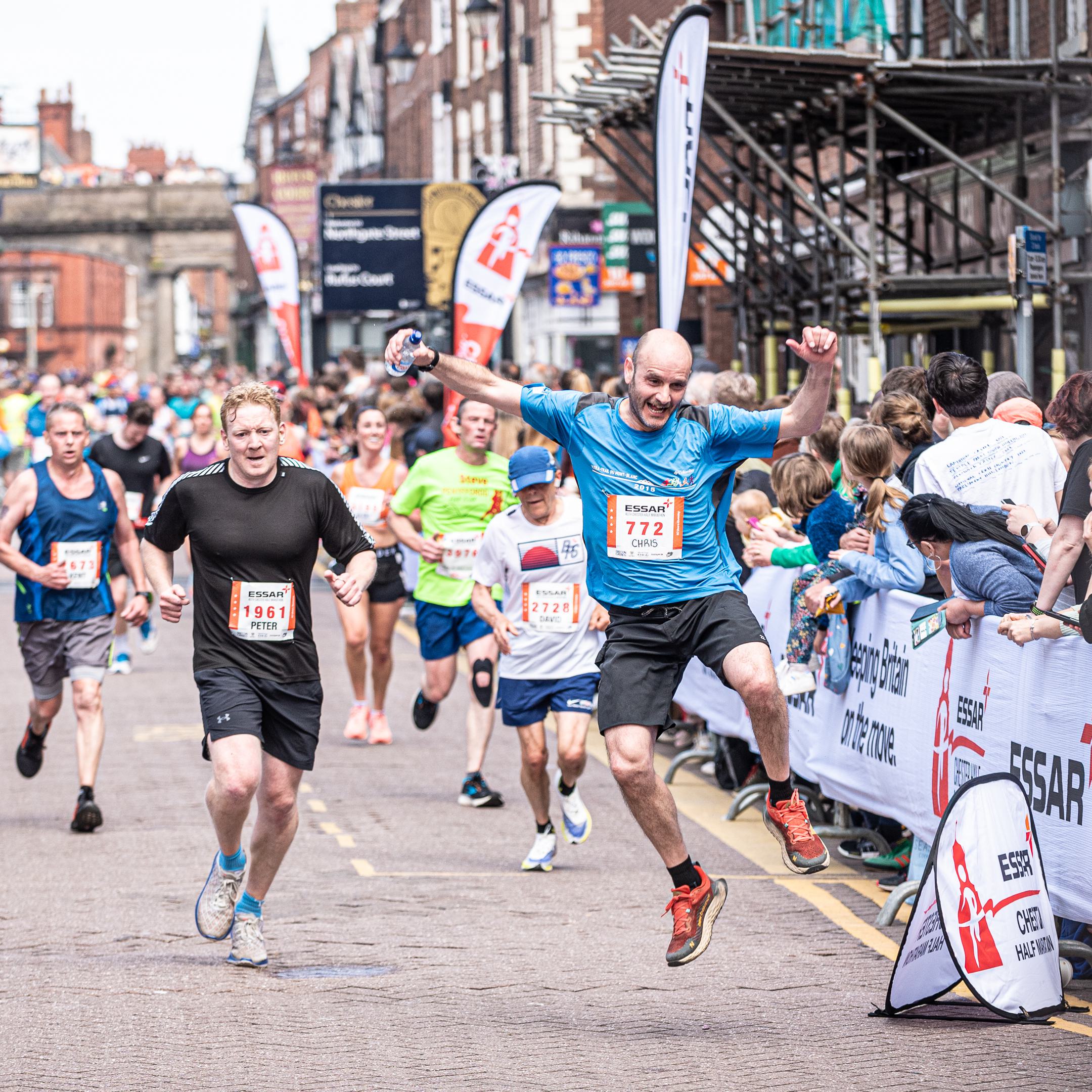 Runners, surrounded by cheering spectators, participate in a lively street race. A man in a blue shirt and black shorts jumps energetically with his arms raised. The scene is vibrant with a mix of runners and enthusiastic onlookers along the course.
