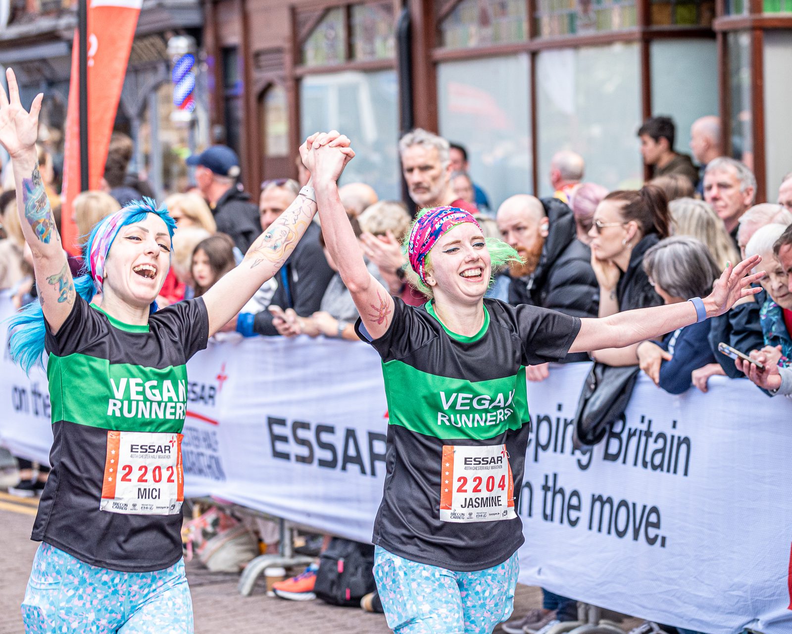 Two runners smile and hold hands triumphantly as they cross the finish line of a race. They wear matching "Vegan Runners" shirts, with bibs labeled "2204" and "2203". Spectators cheer from behind barriers adorned with "Essar" branding and motivational messages.