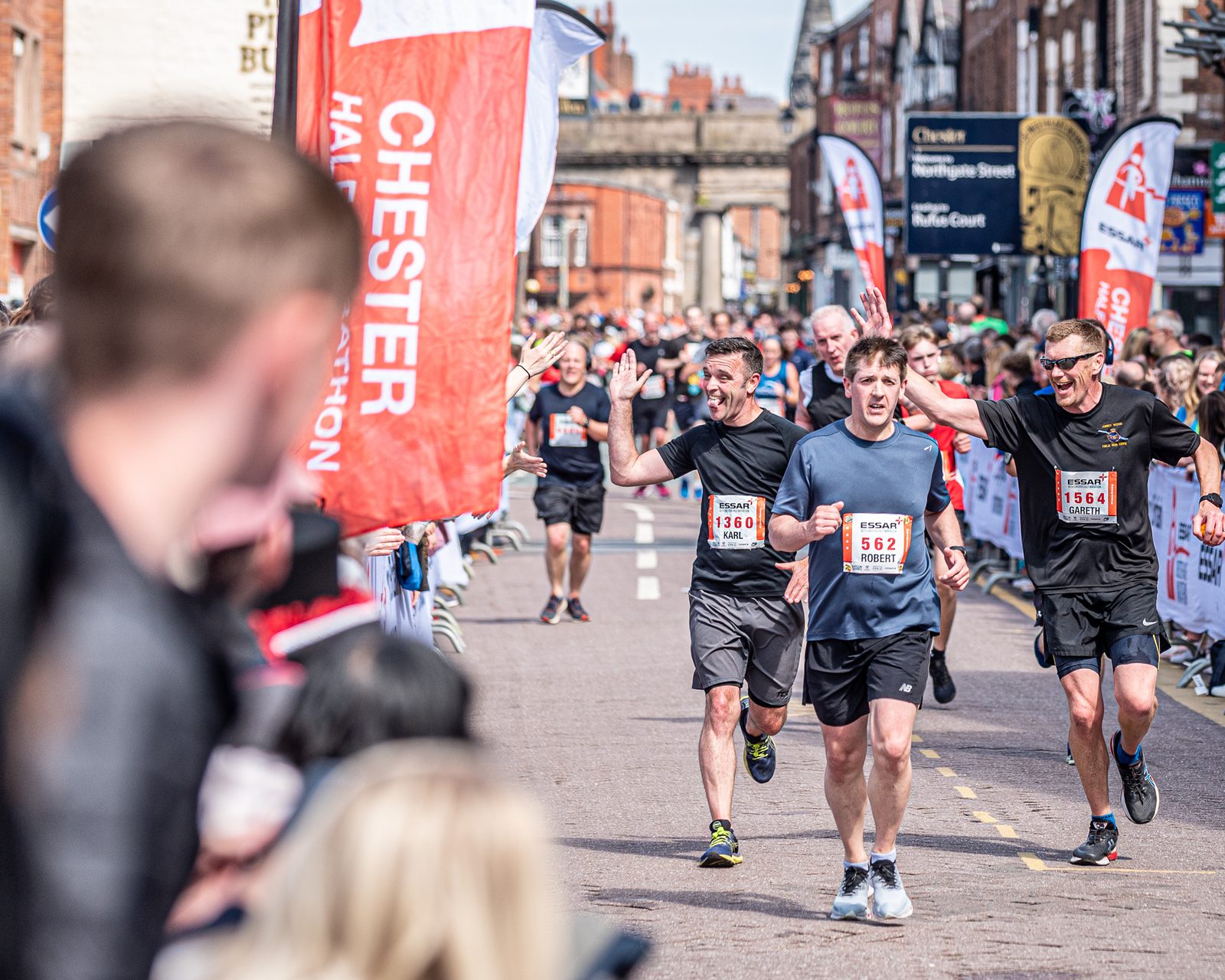 Runners crossing the finish line at the Chester Half Marathon, celebrating with raised hands and smiling. Spectators line the street, cheering and taking photos. Bright banners and flags with event branding are visible in the background.