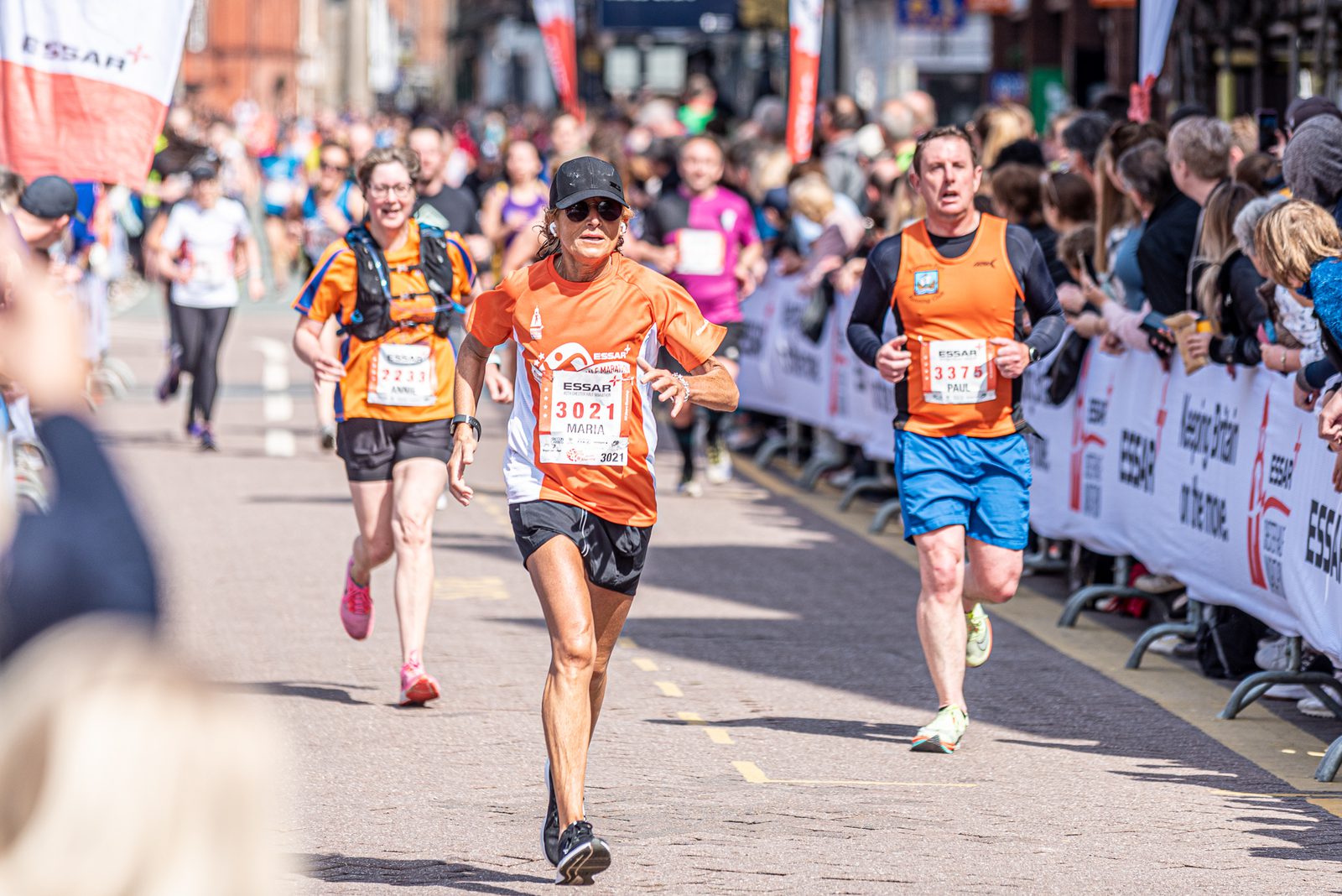 A group of runners participating in a road race, wearing orange shirts with race bibs. They are surrounded by a cheering crowd behind barriers. The athlete in the center is running with determination, while others follow closely behind. Bright and sunny day.