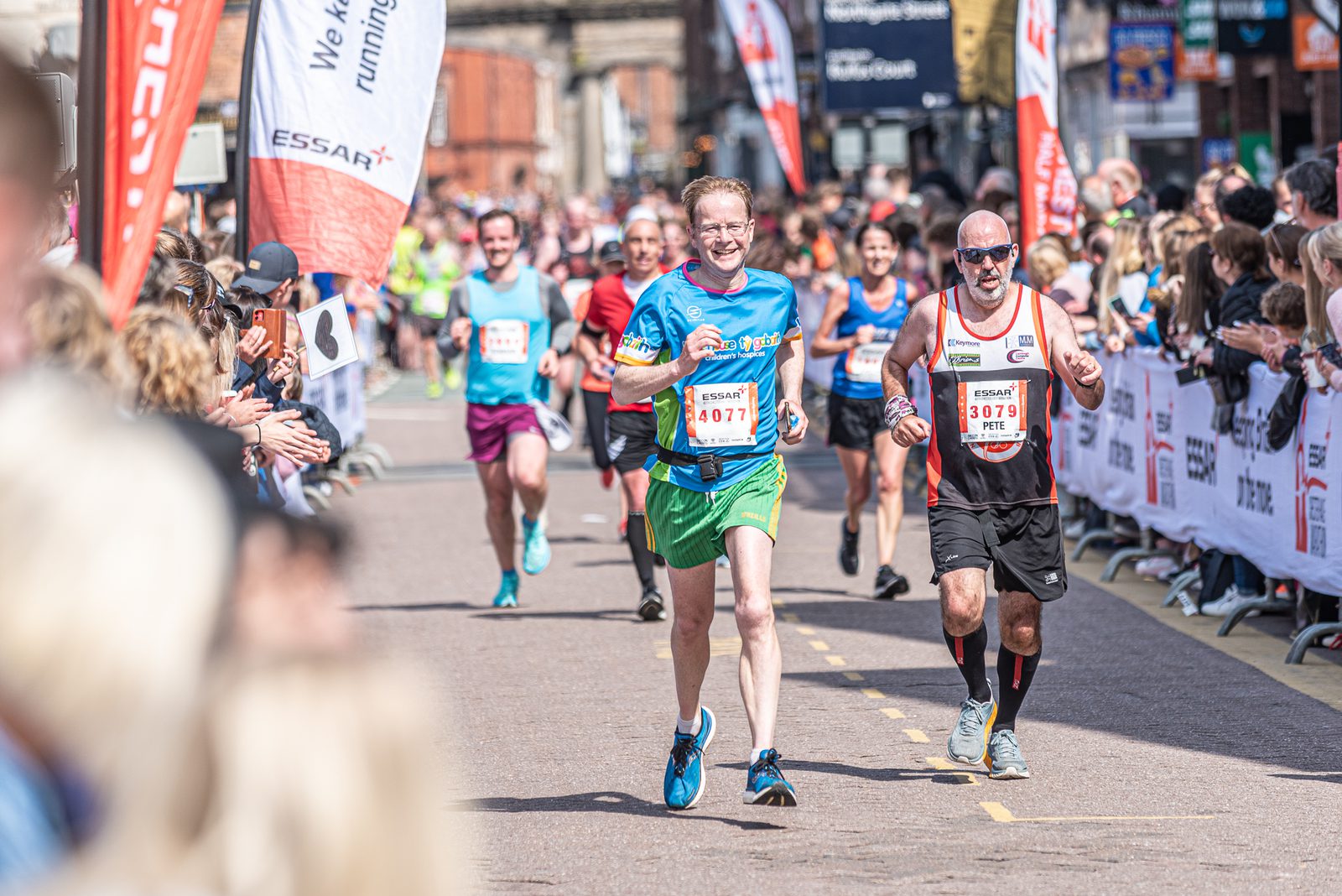 A group of runners in a marathon race, with spectators lining the streets and cheering. Two men in the foreground are running side by side, each wearing a race bib with numbers. The crowd appears enthusiastic, and the atmosphere is lively.