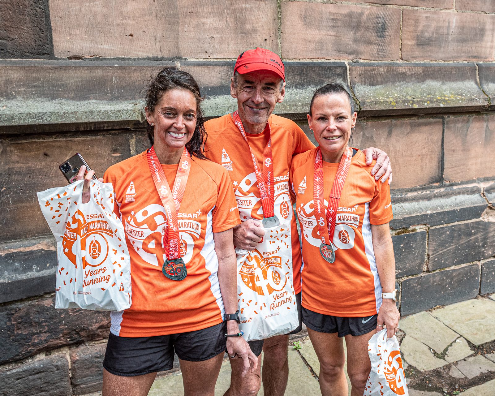Three people in matching orange running shirts and medals stand together holding goody bags. They are smiling, with an old brick wall in the background. The shirts and medals indicate participation in the ESSAR Chester Marathon.