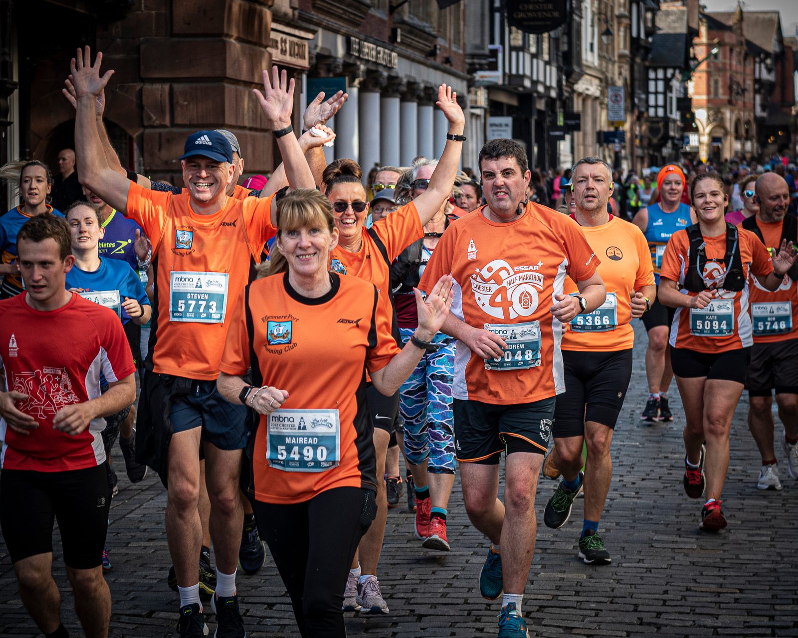A group of people wearing race bibs running on a cobblestone street in a race event. Many are wearing orange shirts, smiling, and raising their arms. Historic brick buildings line the background, with spectators visible. The atmosphere appears lively and enthusiastic.