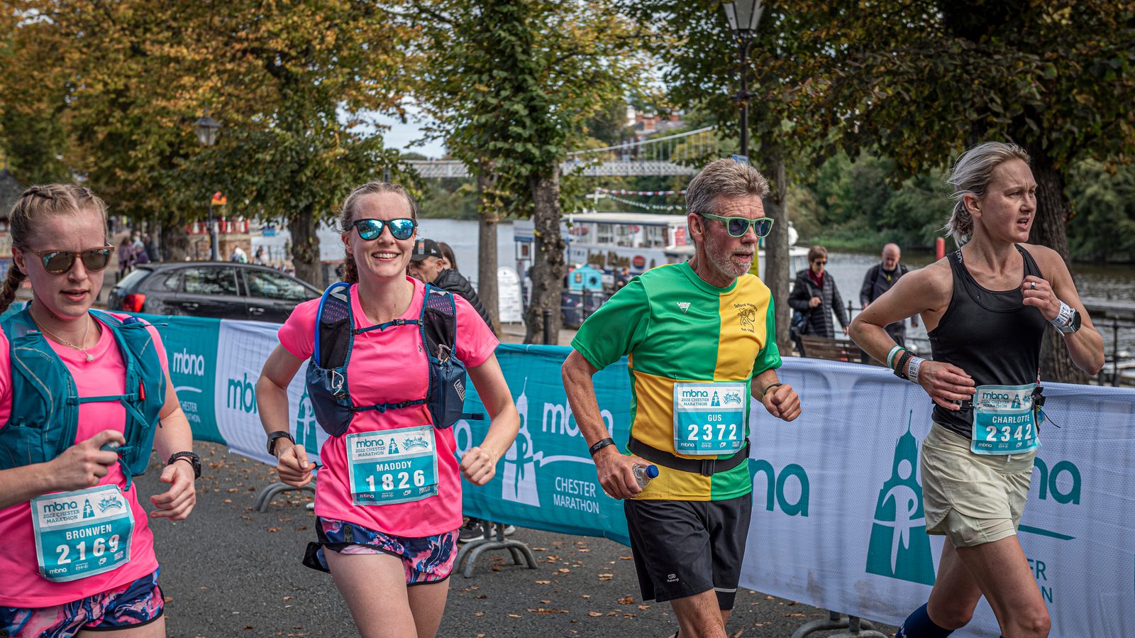 Four runners participate in a marathon event outdoors. They wear race numbers and athletic gear, with a mix of colorful shirts and accessories. The setting is a tree-lined street near a body of water, with onlookers in the background. The atmosphere is energetic.