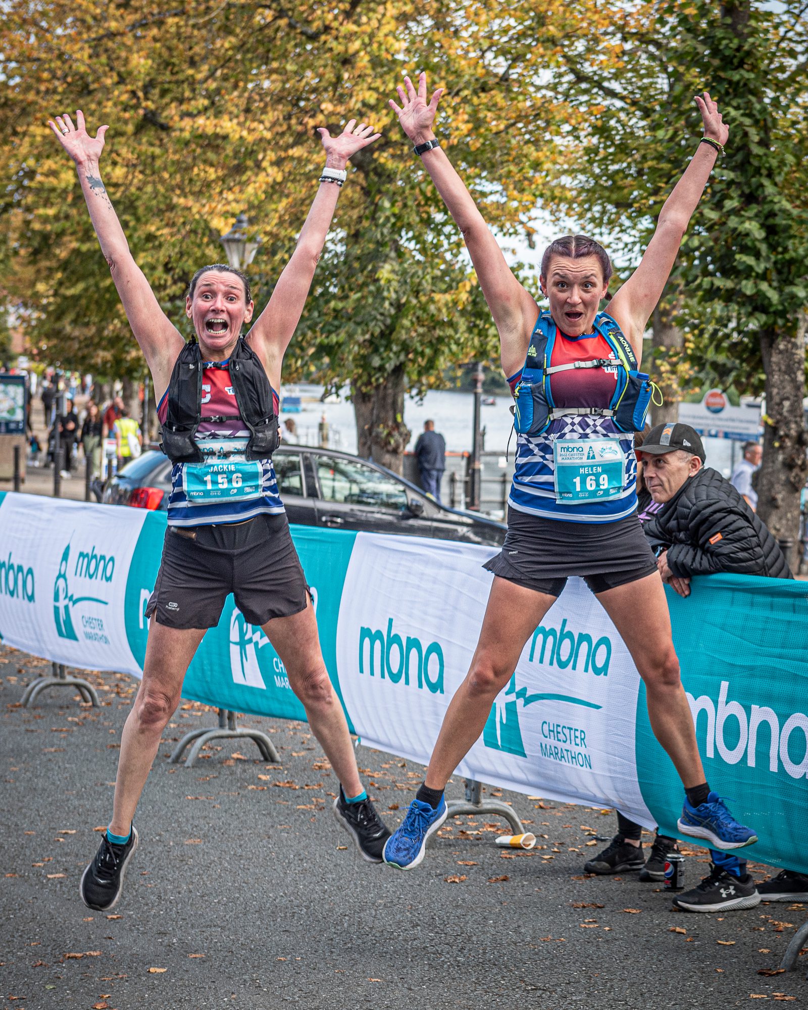 Two people wearing race bibs jump joyfully in the air with arms raised at a marathon event. They are mid-jump near a race barrier with "mbna" logos and a sign that reads "Chester Marathon." Trees with autumn leaves are visible in the background.