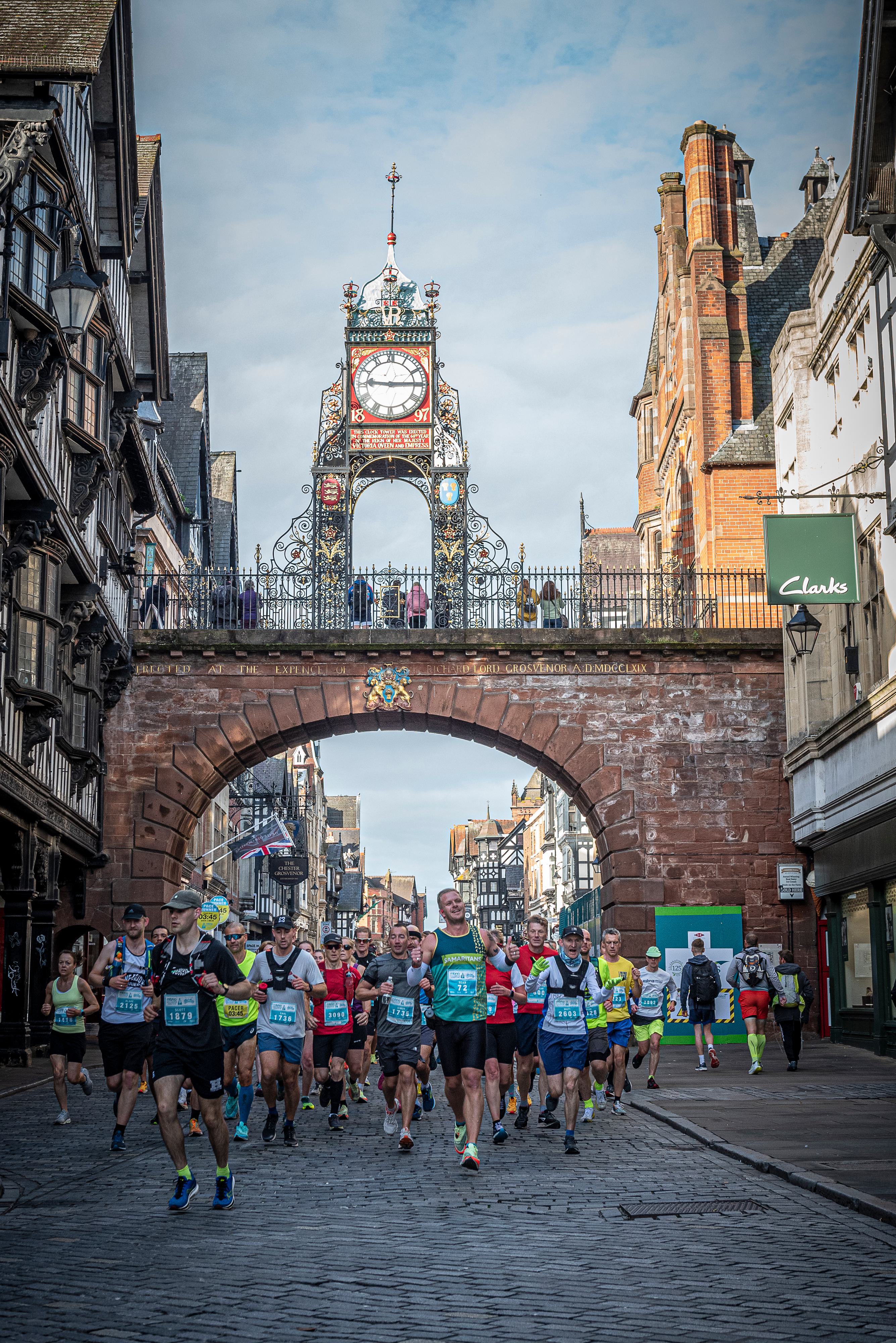 A group of runners competes in a race on a cobblestone street in front of the Eastgate Clock in Chester, UK. Historic buildings line the street on both sides. The runners wear various colored athletic gear under a cloudy sky. Spectators watch from the walkway above.