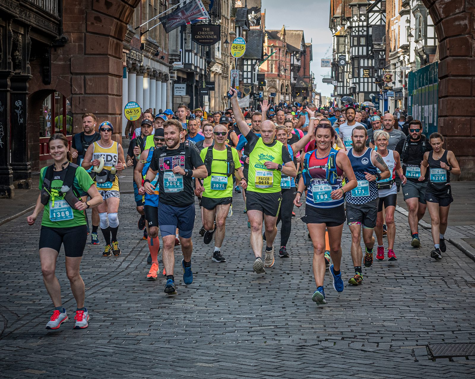 A diverse group of runners participating in a road race, running through a historic street lined with old buildings. The lead runner, with a green tank top and black shorts, raises his arm, smiling. The runners wear race bibs, and some carry bright balloons.