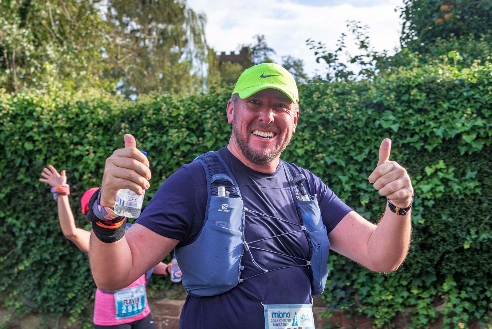 A man in a bright green cap smiles and gives two thumbs up while participating in a marathon. He is wearing a blue vest with hydration packs and a navy shirt. There are other runners and greenery in the background.