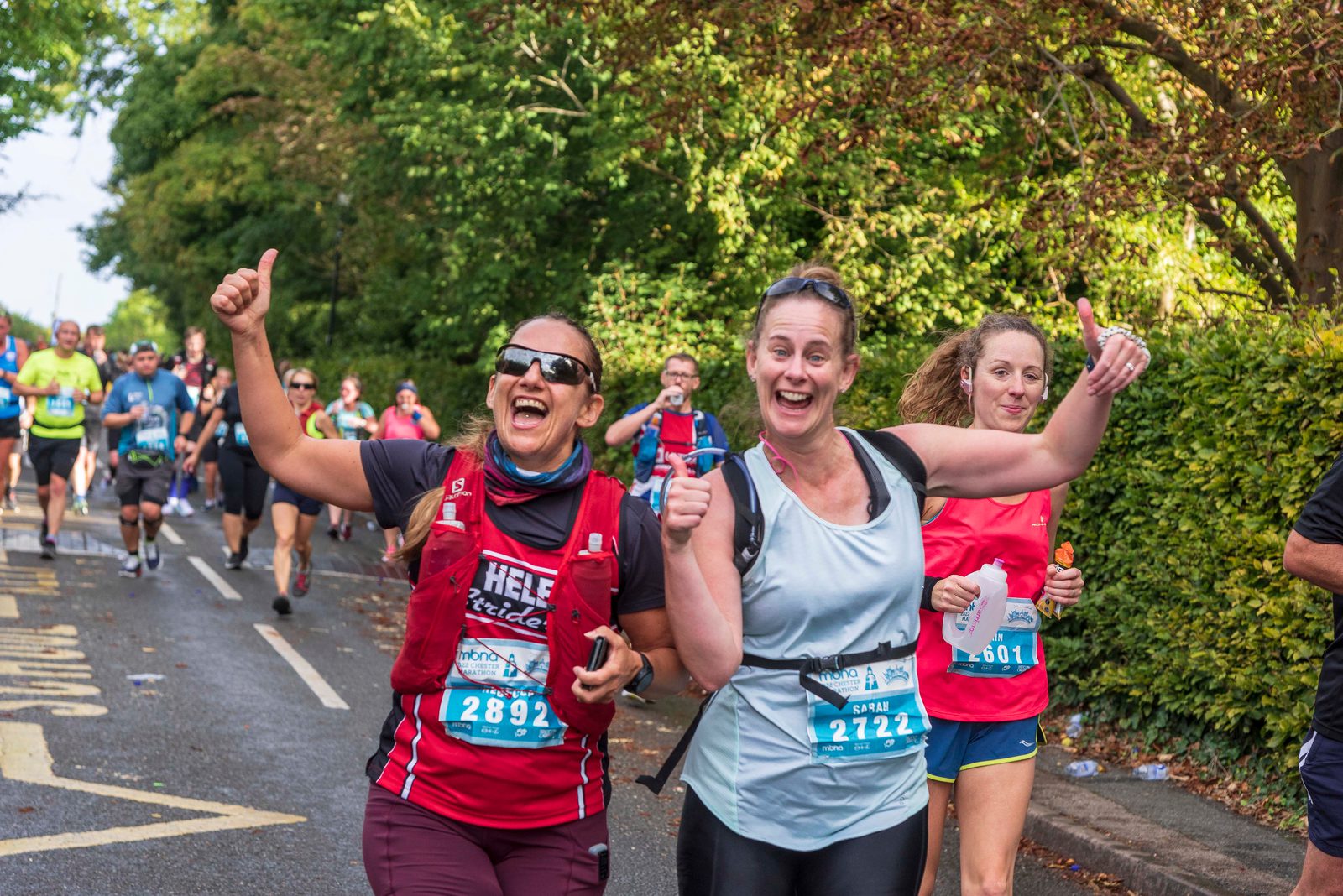 A group of people participate in a marathon. Two women in the foreground, wearing race bibs 2892 and 2722, are smiling and giving thumbs up. Participants behind them run along a tree-lined street on a sunny day.