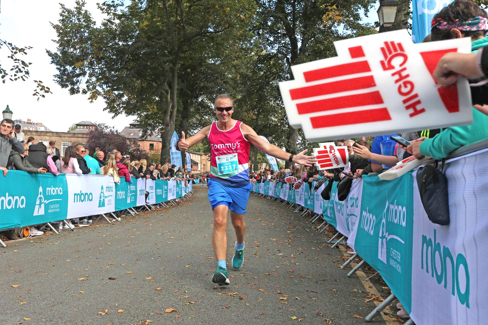 A marathon runner in blue shorts and a pink tank top high-fives spectators holding foam hands as he approaches the finish line. The sunny outdoor event has many cheering attendees and is decorated with sponsor banners.