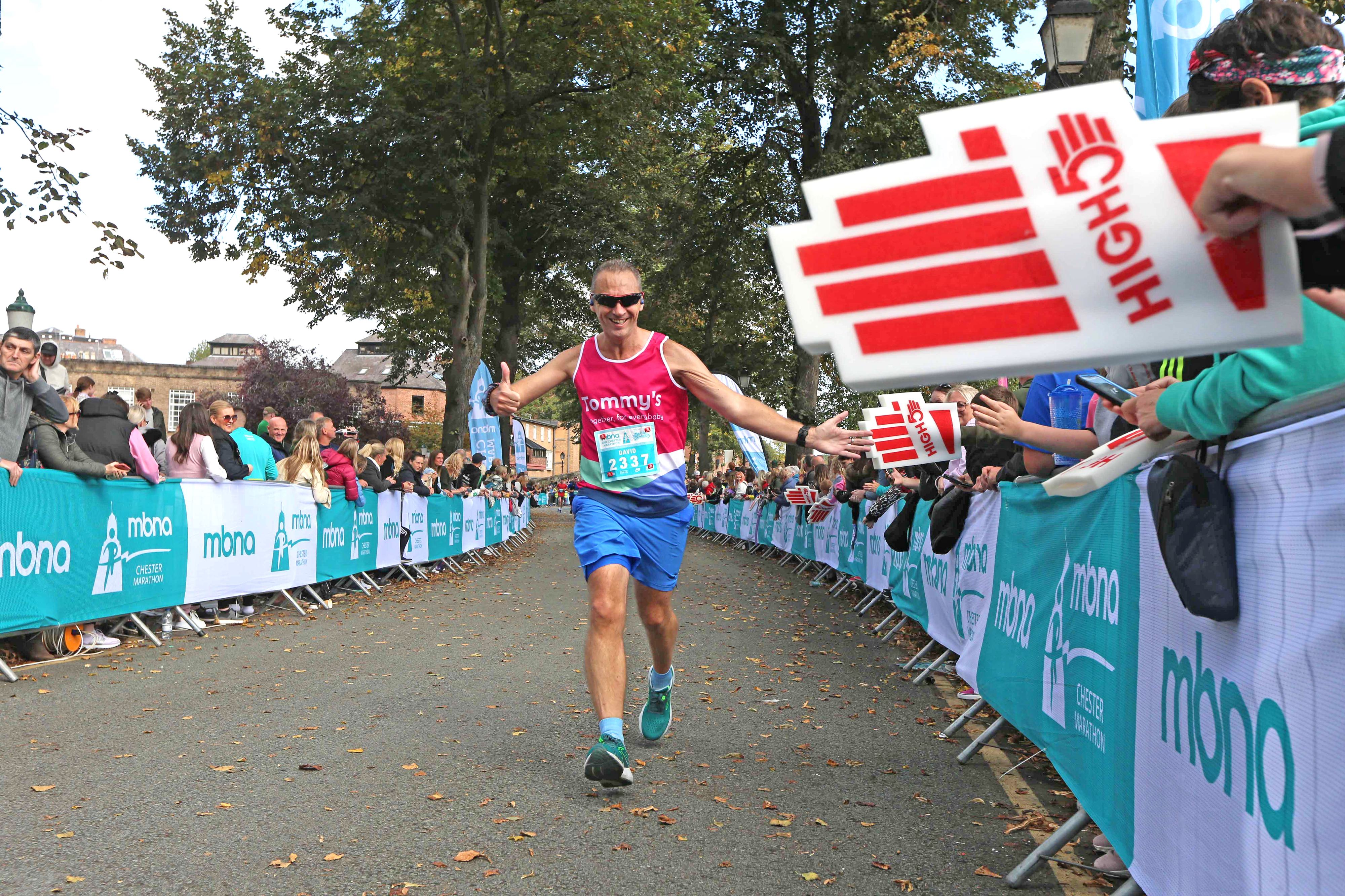 A marathon runner in blue shorts and a pink tank top high-fives spectators holding foam hands as he approaches the finish line. The sunny outdoor event has many cheering attendees and is decorated with sponsor banners.