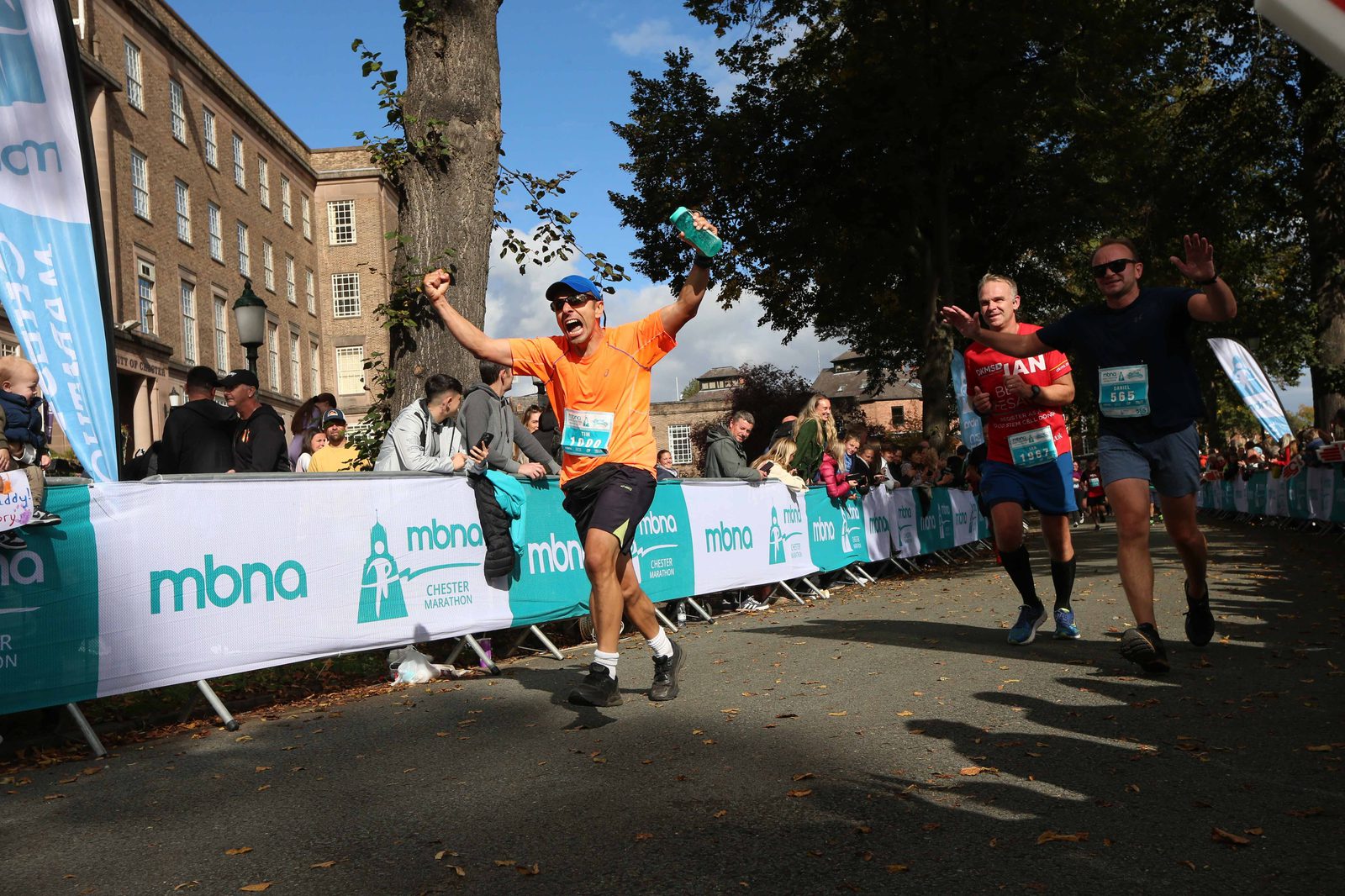 A runner in an orange shirt triumphantly raises his arms as he crosses the finish line during the MBNA Chester Marathon. Other runners are seen in the background, and spectators line the path behind a white barricade with MBNA branding.
