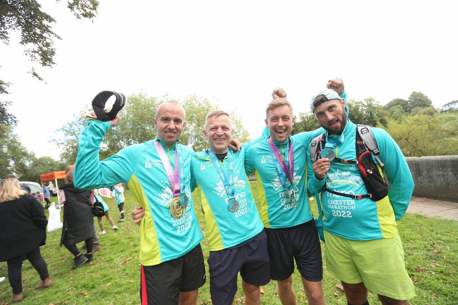 Four men stand together outdoors, celebrating and smiling. They are wearing matching teal athletic shirts with "2022" and "Chester Marathon" printed on them, and each has a medal around their neck. One man is holding a cap in the air, and trees and other people are in the background.
