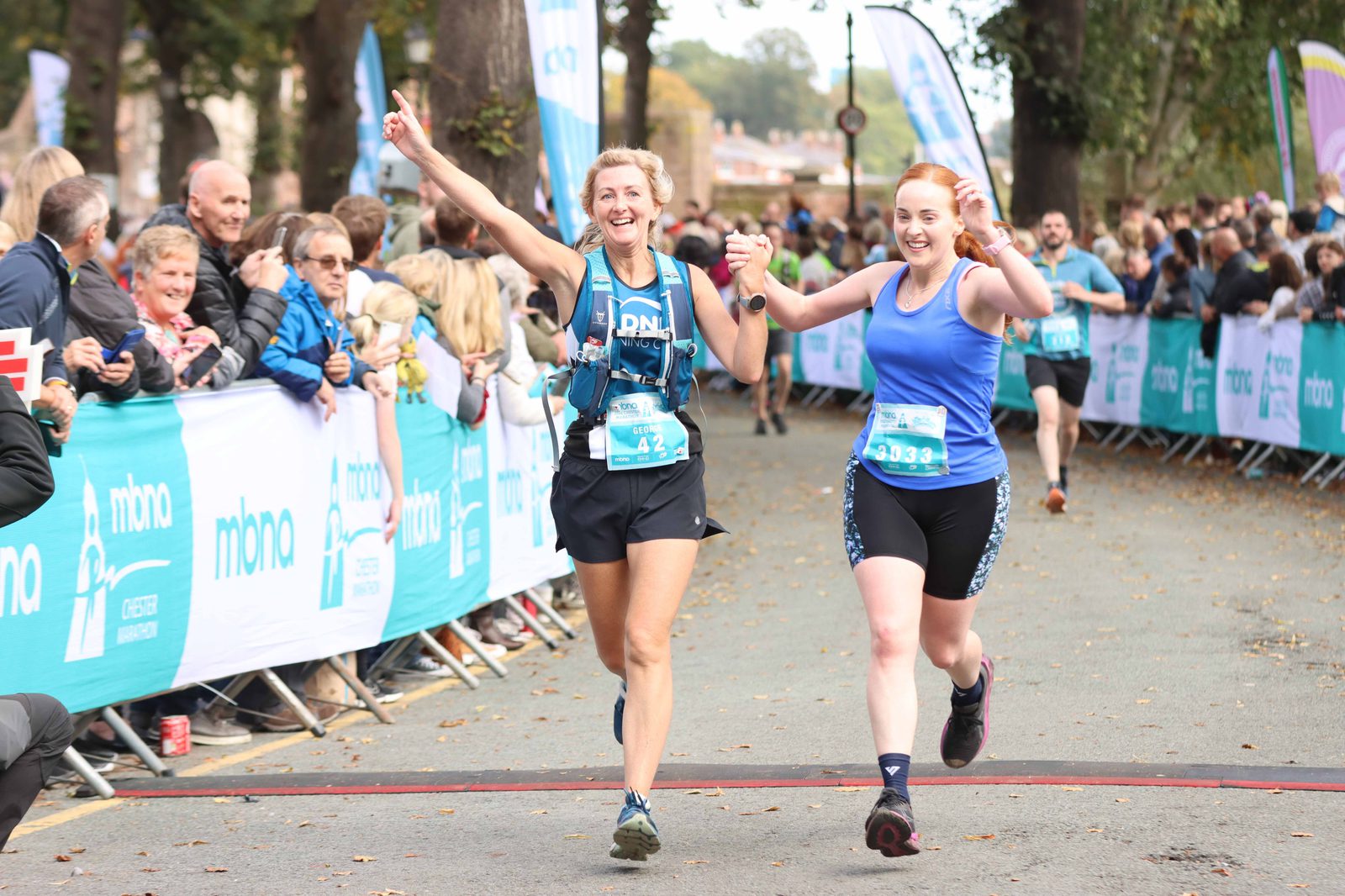 Two women joyfully cross the finish line of a race, holding hands and smiling. Both wear athletic gear, bib numbers visible. The road is lined with cheering spectators behind barriers. Trees and banners add to the lively atmosphere.