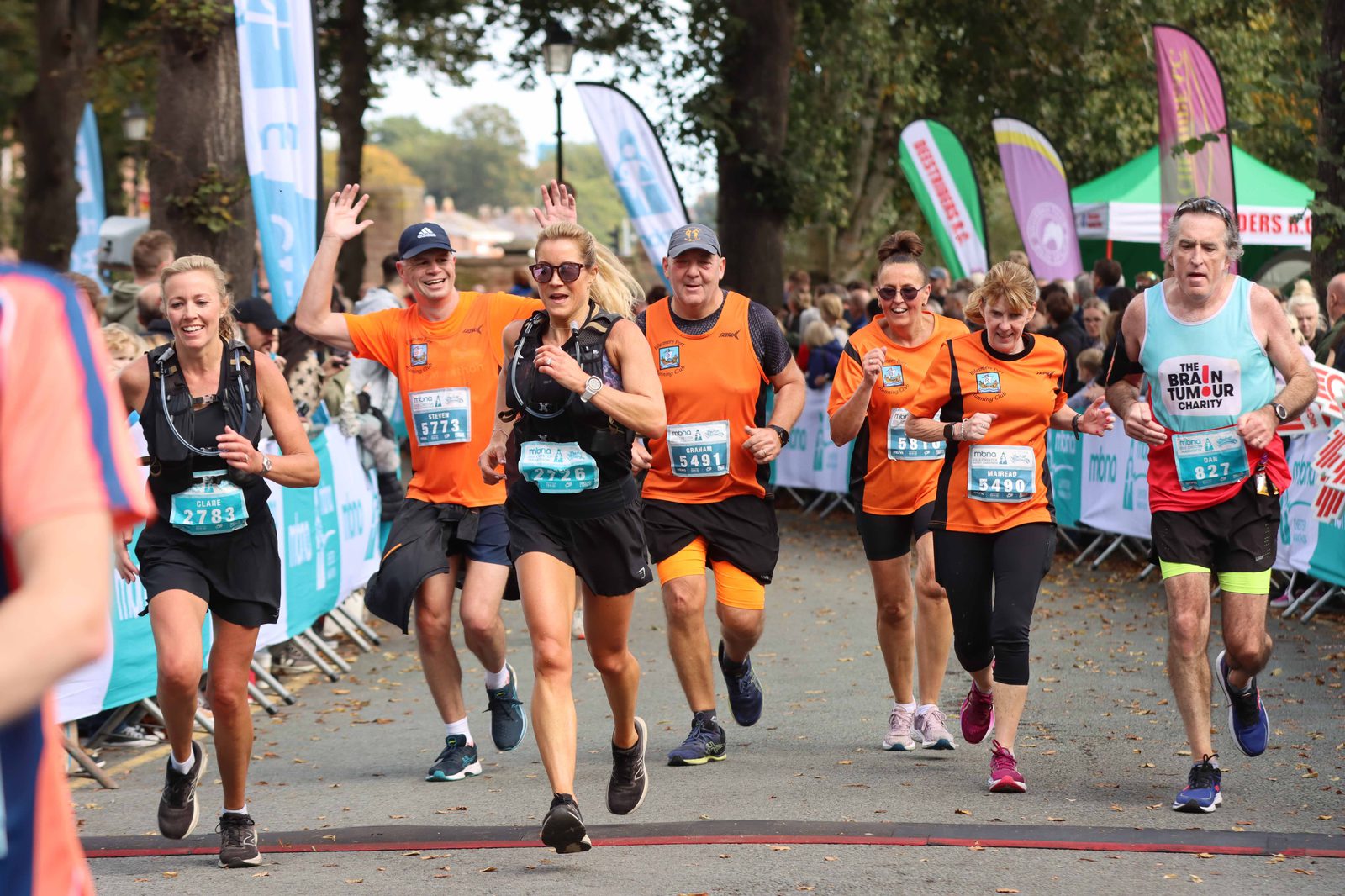 A group of runners, some in orange shirts and others in black, are participating in a race on a tree-lined street. They appear excited and are smiling, with some waving at the camera. A person in the foreground is wearing a "brain tumor" awareness shirt. Bystanders and colorful banners line the course.