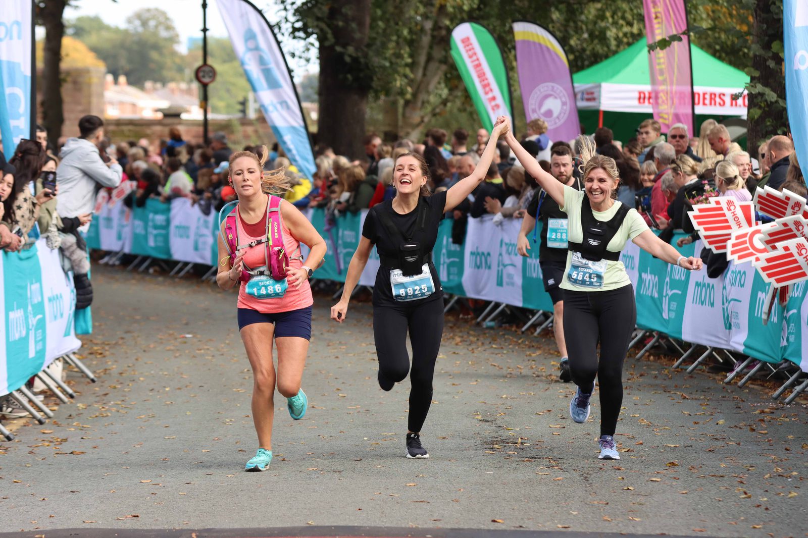 Three women, wearing running gear and race bibs, joyfully cross the race finish line holding hands. They are surrounded by cheering spectators and banners. The atmosphere is lively and celebratory with trees and a blue sky in the background.