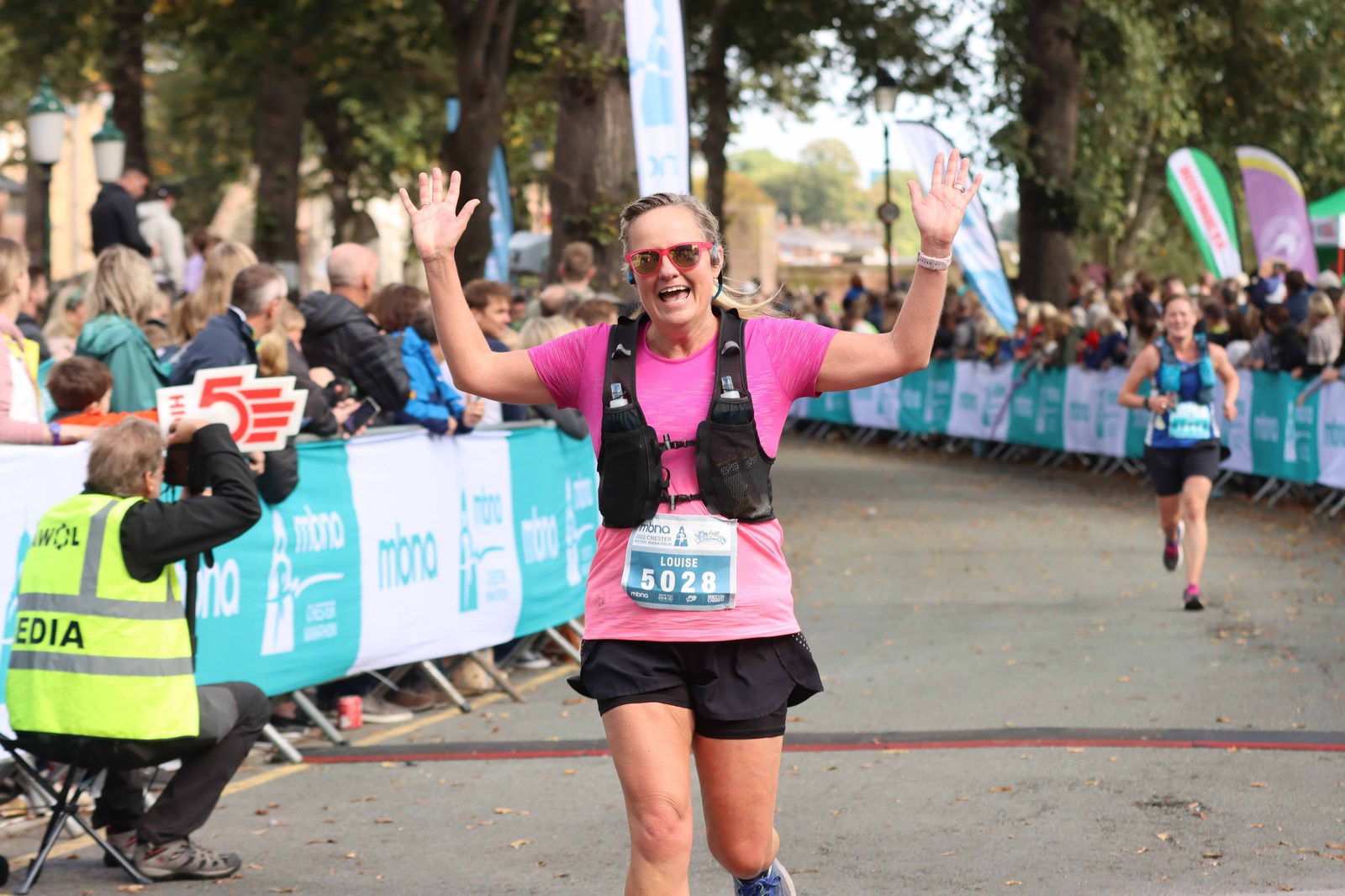 A smiling runner with raised arms crosses the finish line in a race. She is wearing a pink shirt, black shorts, and a race bib numbered 5028. Spectators line the path behind barricades, and a man with a media vest is on the side. Trees line the background.