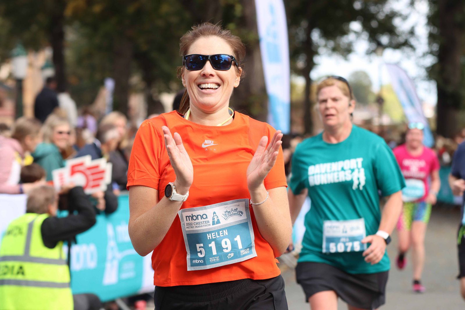 A woman wearing an orange shirt, black shorts, and sunglasses smiles and claps while running in a race. She has a race bib with the name "Helen" and the number "5191." Other runners and spectators are visible in the background, including one wearing a teal "Davenport Runners" shirt.