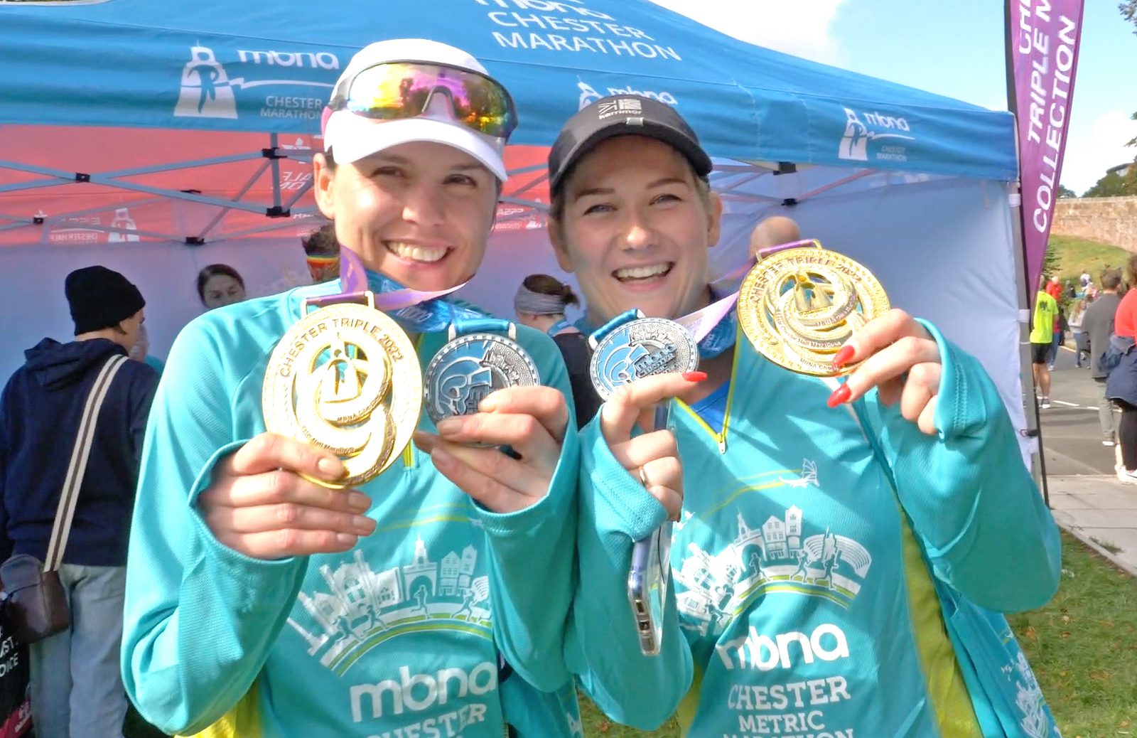 Two smiling women wearing matching blue and green athletic shirts and caps proudly hold up gold, silver, and bronze medals. They stand in front of a tent with "mbna Chester Metric" visible on it, indicating they participated in the Chester Marathon event.
