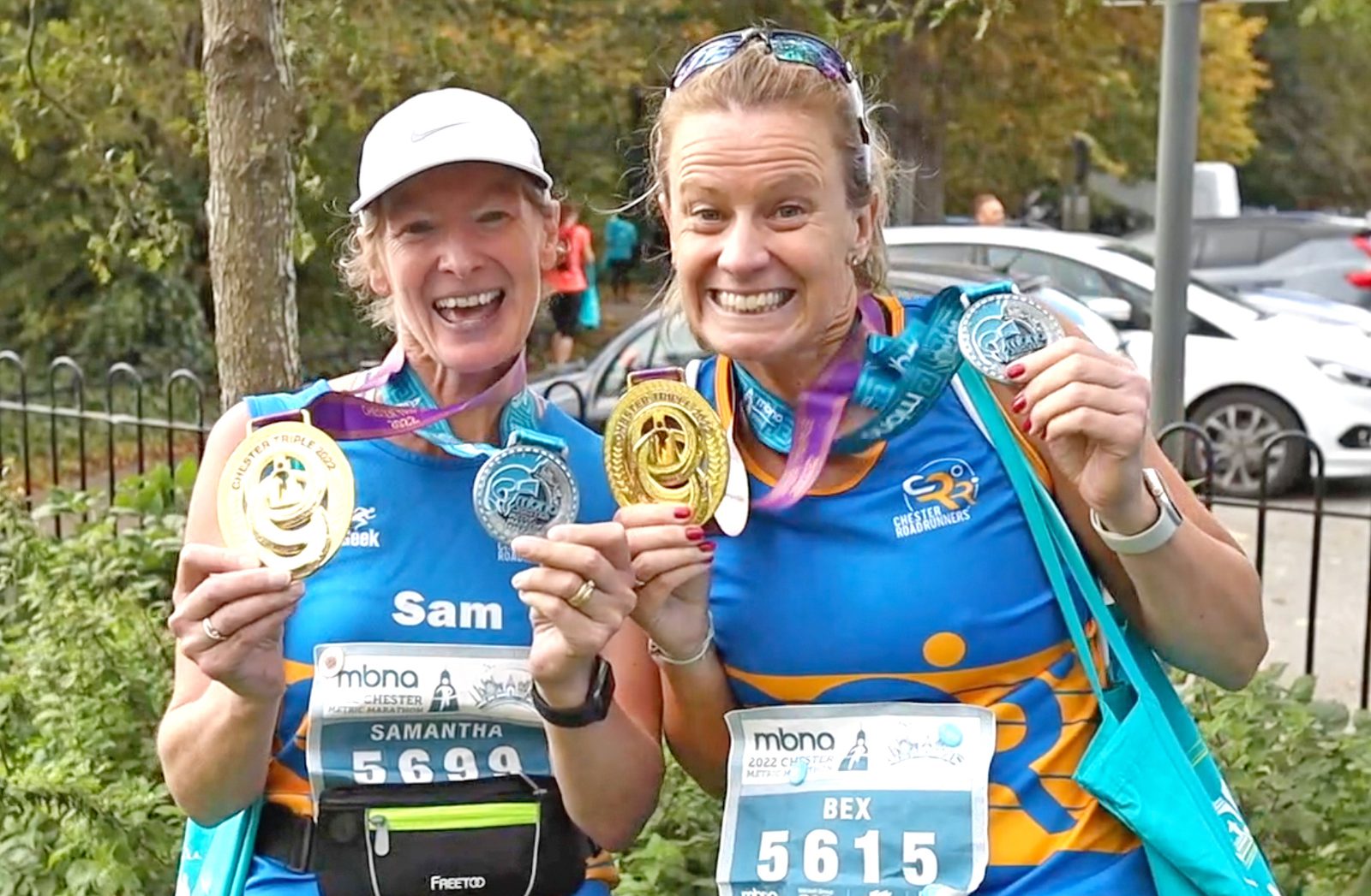 Two cheerful runners, Sam and Bex, wearing blue athletic gear, proudly display their medals after a race. Both hold multiple medals and smile at the camera. Sam’s bib number is 5699, and Bex’s is 5615. Trees and parked cars are visible in the background.