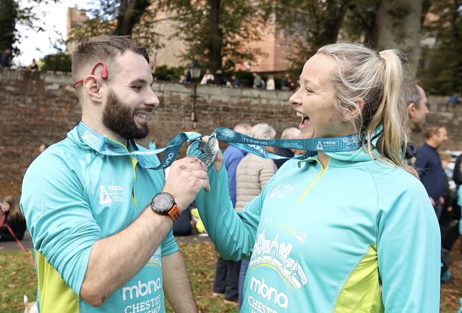 A man and woman, both in light blue athletic tops, stand outdoors, laughing and holding medals around their necks. They appear joyful, suggesting they've just completed a race. People and trees are in the background.