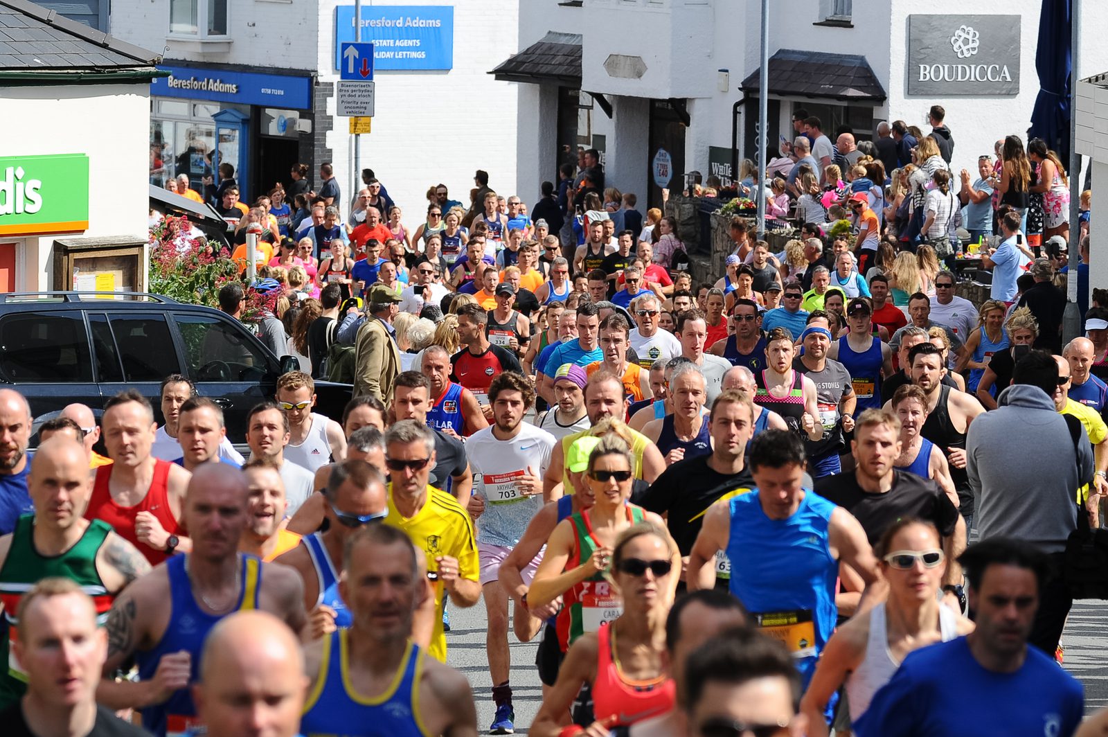 A large group of runners participating in a race through a town. The street is lined with spectators on both sides, and various shops and buildings are visible in the background. The runners wear a mix of brightly colored athletic clothing and appear to be in mid-stride.