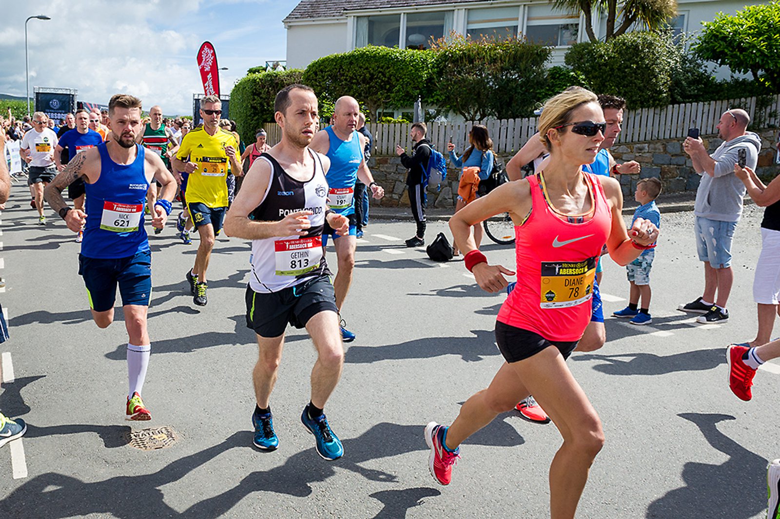 A group of runners, wearing numbered bibs and athletic gear, participate in a road race. The leading runner is a woman in a pink tank top and sunglasses. Spectators line the sidewalks, some clapping and taking photos. Trees and a building are in the background.