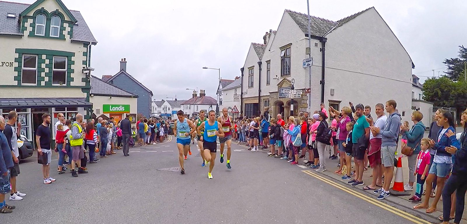 A group of runners participates in a race on a street lined with spectators. The buildings on either side include a green and white shop and a white building with a sloped roof. The crowd cheers as the runners pass by, and the sky is overcast.