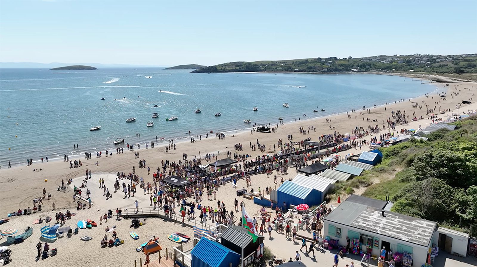 Aerial view of a lively beach scene on a sunny day. Crowds of people are gathered on the sandy beach and in the water. Several boats are visible near the shore. There are colorful beach huts, umbrellas, and buildings along the coastline with rolling green hills in the background.