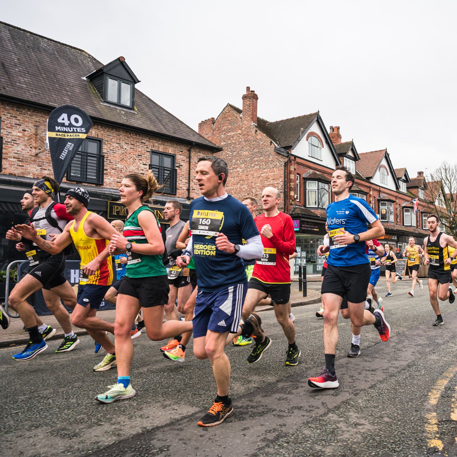 A group of runners participating in a road race passes by brick buildings and spectators. One runner is holding a black-and-white sign that reads "40 MINUTES." The runners wear colorful athletic gear, numbered bibs, and appear focused on their performance.