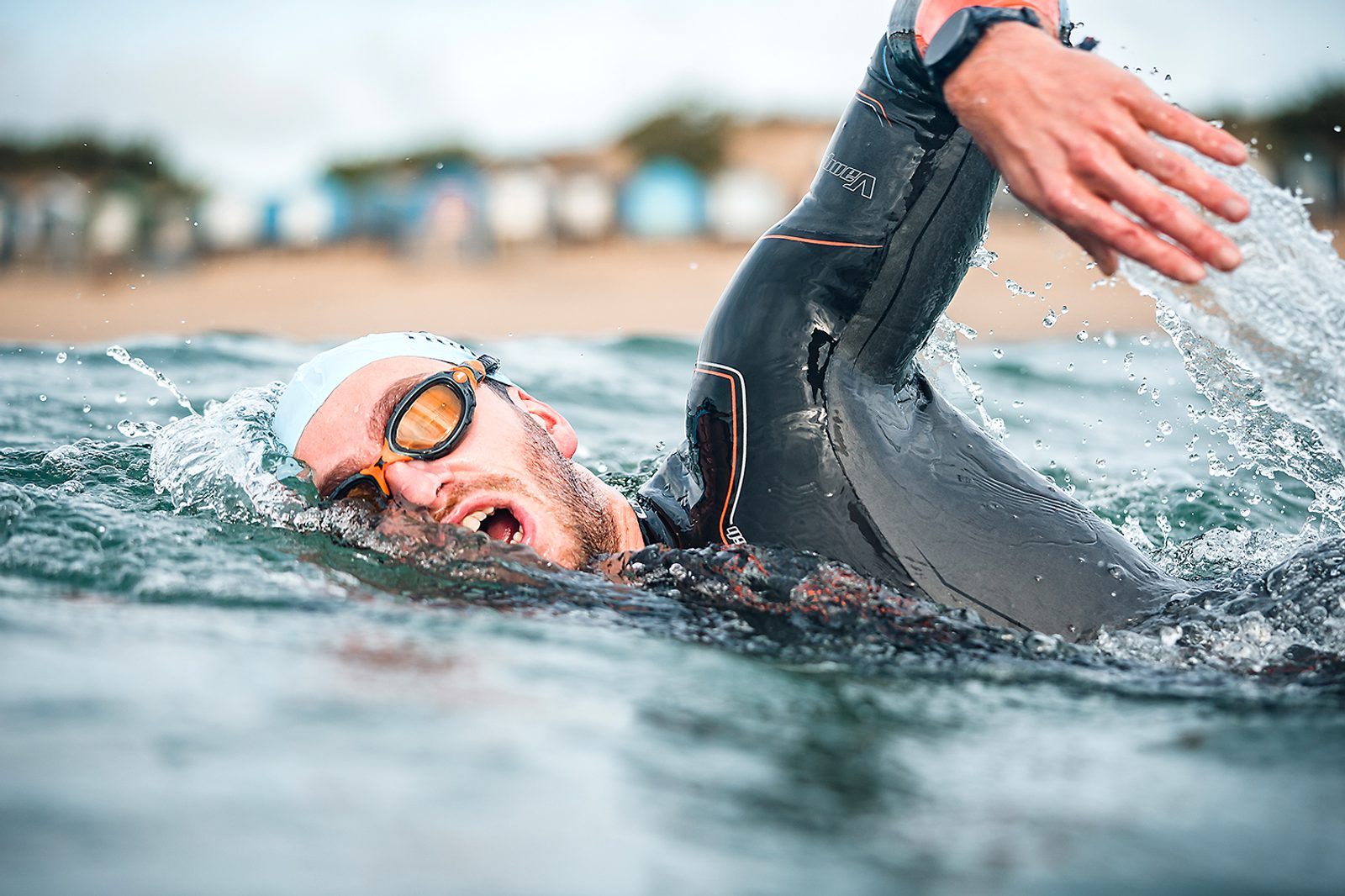 A swimmer wearing a wetsuit, goggles, and a swim cap is captured mid-stroke while swimming in open water. The water splashes around him, and a shoreline with blurred buildings is visible in the background.