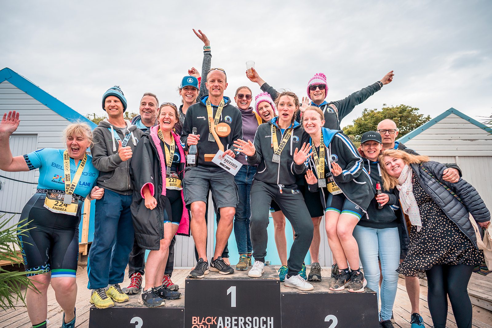 A group of cheerful people stands on a podium celebrating at an outdoor event. Some participants wear medals and athletic gear, while others are in casual clothing. They pose excitedly with smiles, waving their hands in the air. Colorful beach huts are in the background.