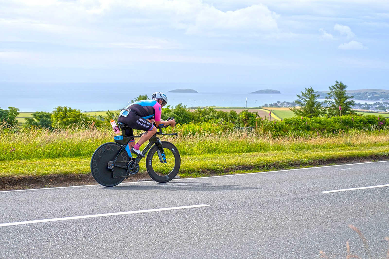 A cyclist in racing gear rides a time trial bike on a rural road with lush greenery and ocean views in the distance. The rider is leaning forward, concentrating on speed, with a helmet and aerodynamic wheels visible. The sky is partly cloudy.